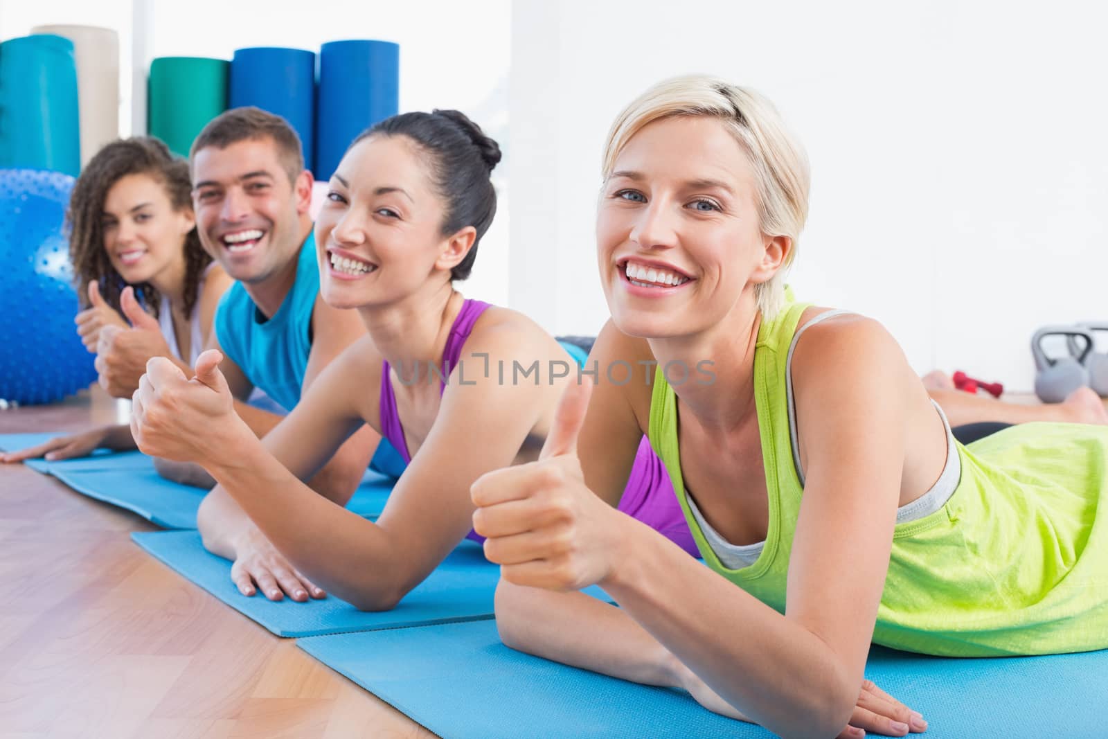 Portrait of happy woman with friends gesturing thumbs up while lying on exercise mats at gym