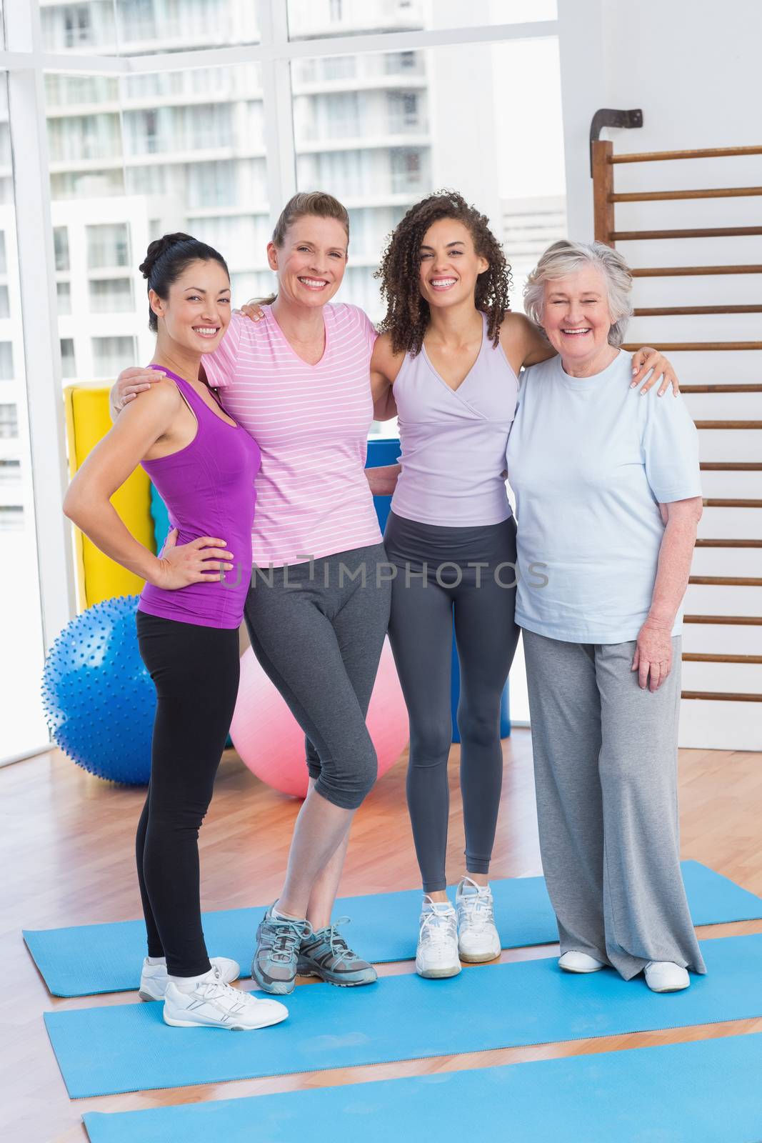Full length portrait of happy women standing arms around in gym