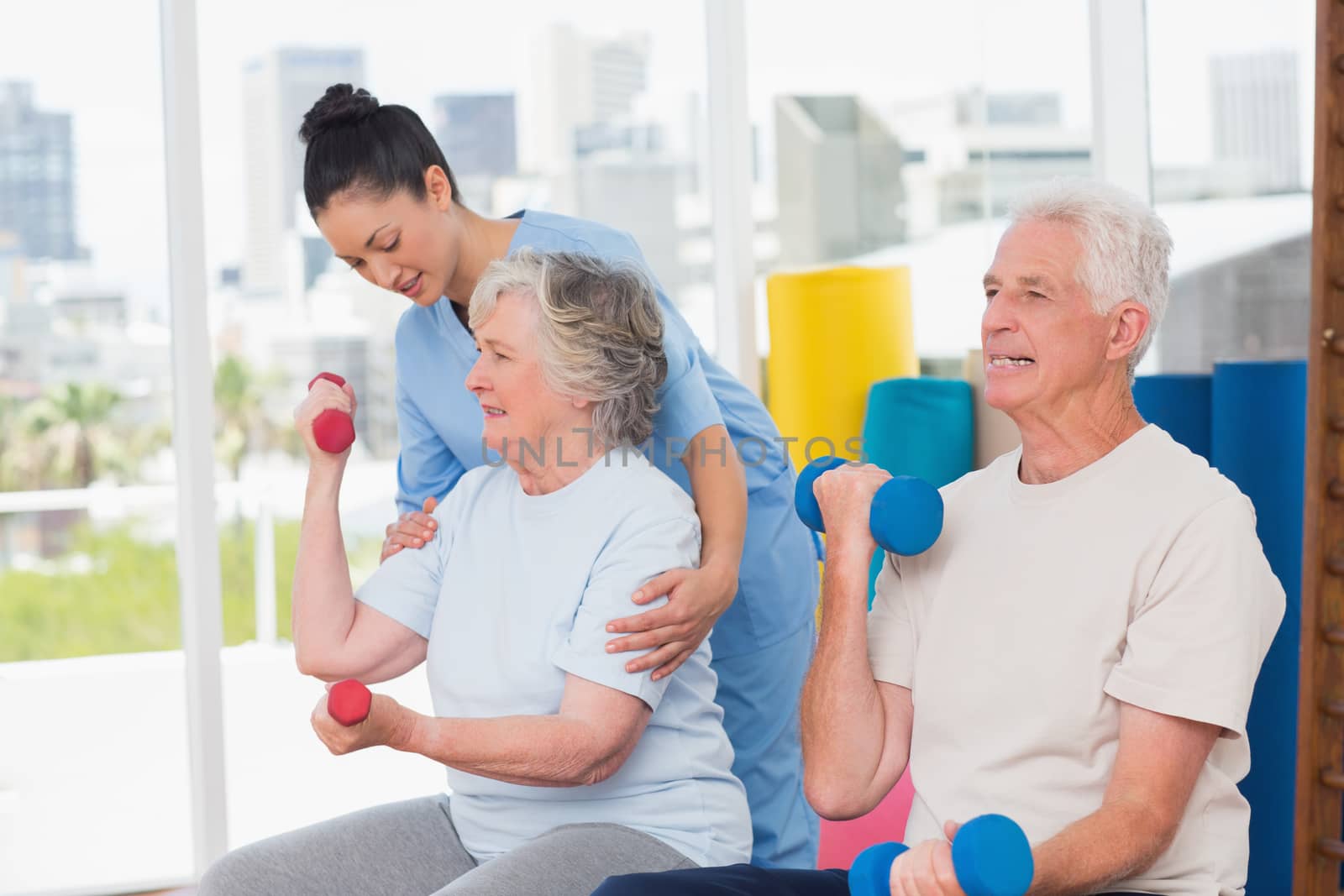 Young female instructor assisting senior woman in lifting dumbbells by man at gym