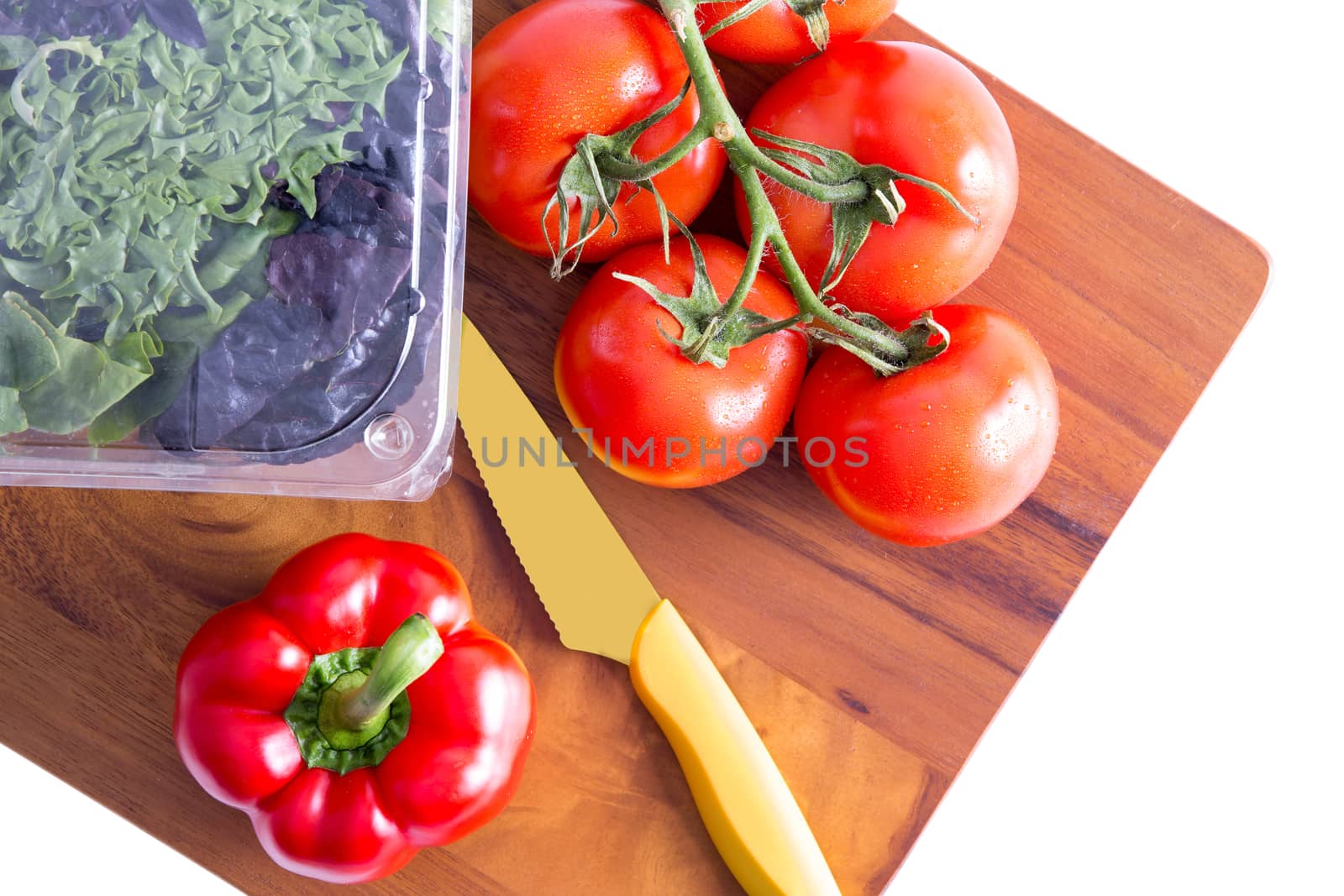 Healthy fresh salad ingredients on a chopping board with a sharp paring knife with three varieties of lettuve in a punnet and vine tomatoes with red bell pepper alongside, isolated on white