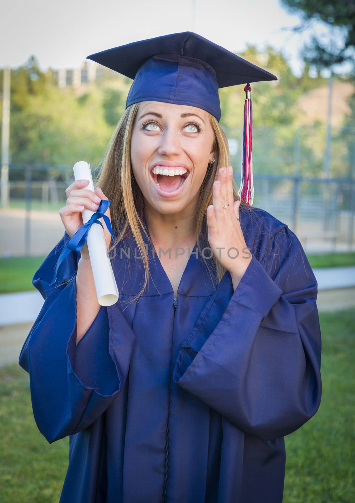 Excited and Expressive Young Woman Holding Diploma in Cap and Gown Outdoors.