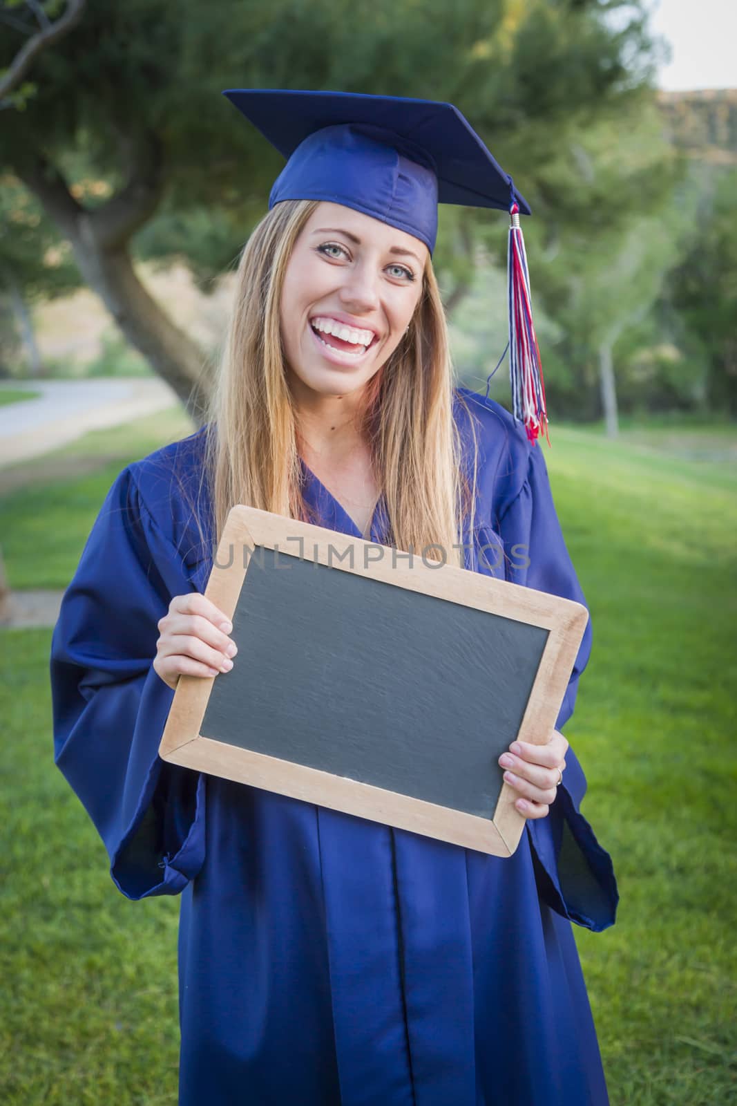 Excited Young Woman Holding Diploma and Blank Chalkboard Wearing Cap and Gown Outdoors.