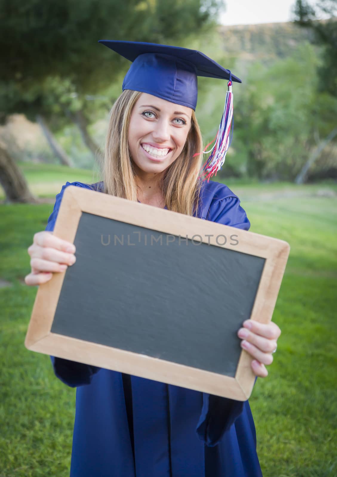 Excited Young Woman Holding Diploma and Blank Chalkboard Wearing Cap and Gown Outdoors.