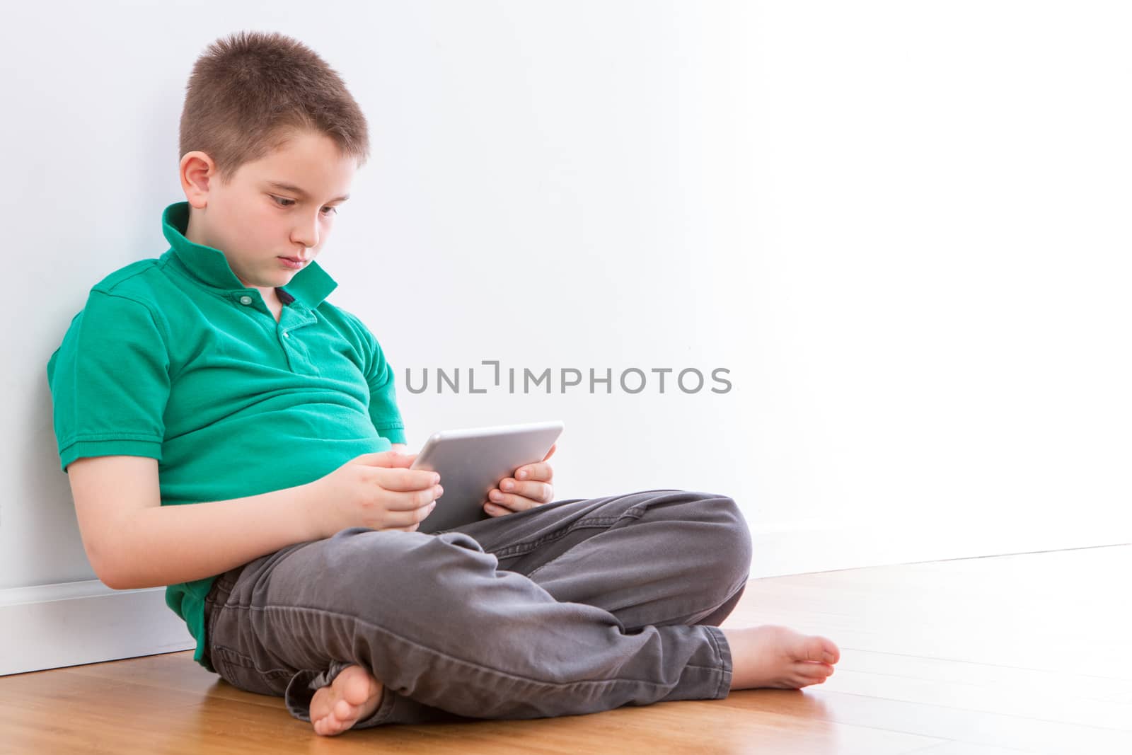 Close up Serious Handsome Young Boy Playing at his Tablet Computer, Sitting on the Floor Inside the House and Leaning on the Wall.