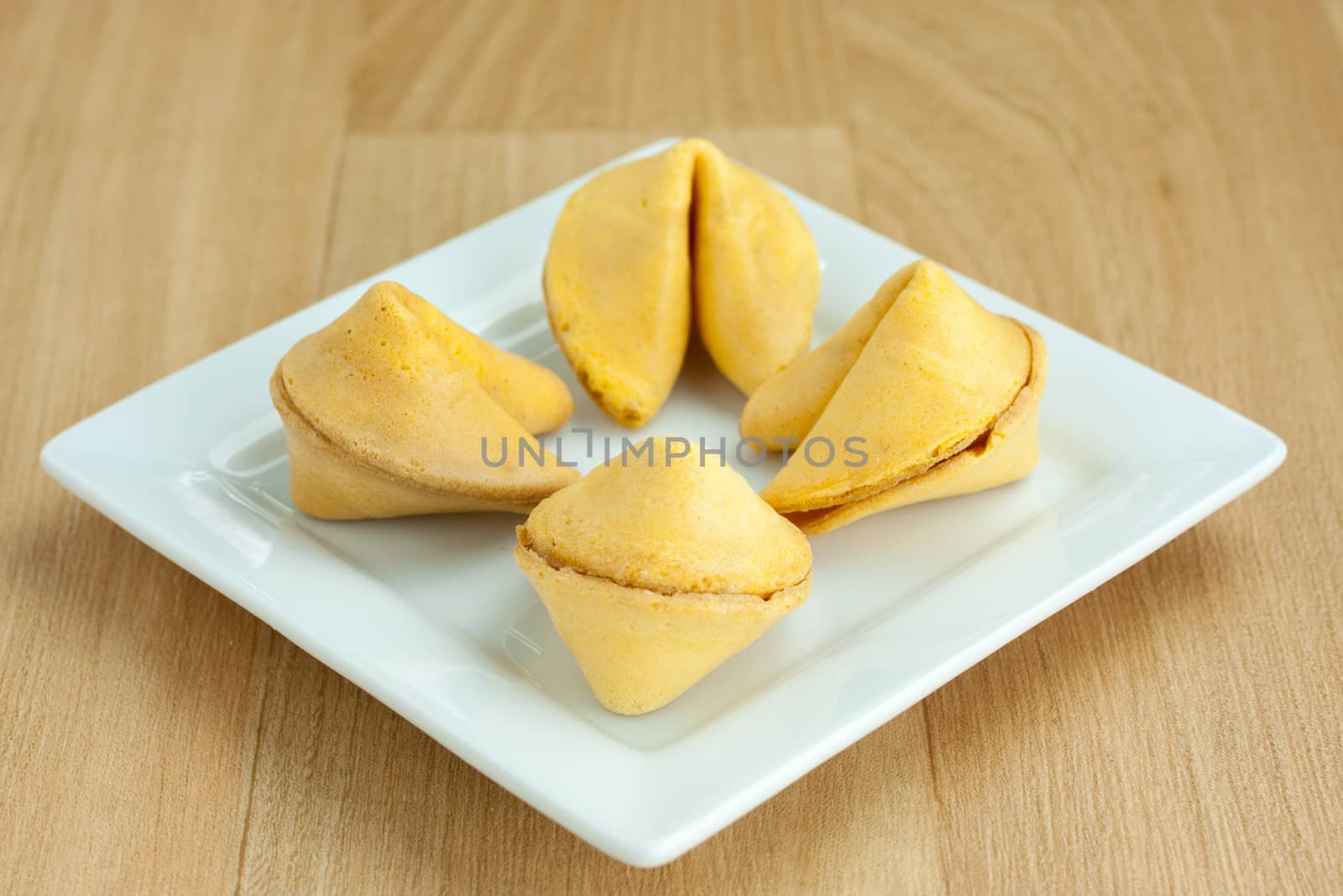 Four fortune cookies on a square white plate and sitting on a wooden table.