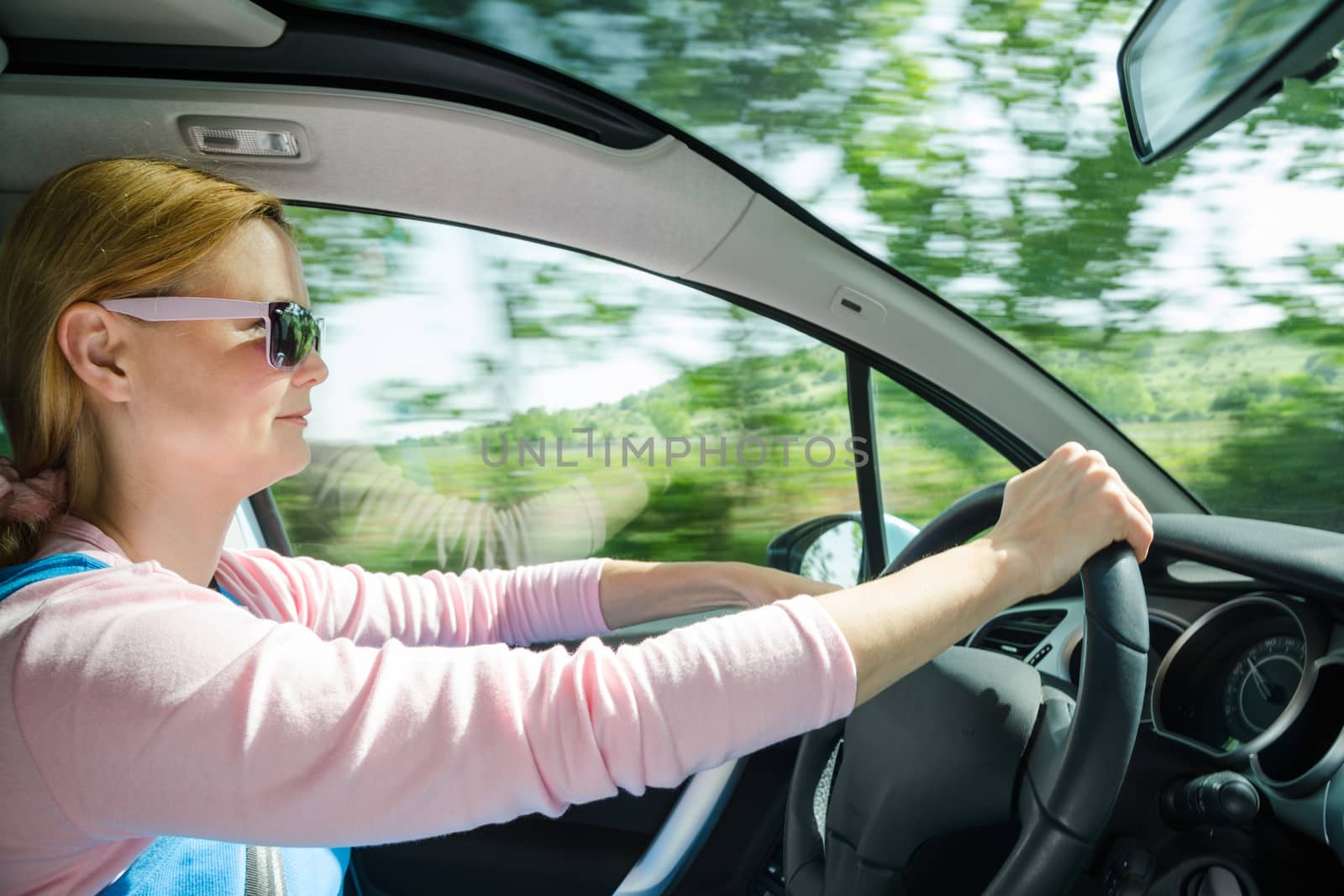 Smiling beautiful woman in sunglasses driving at high speed car with panoramic windshield. Internal stock photo with low shutter speed and blurred in motion natural background.