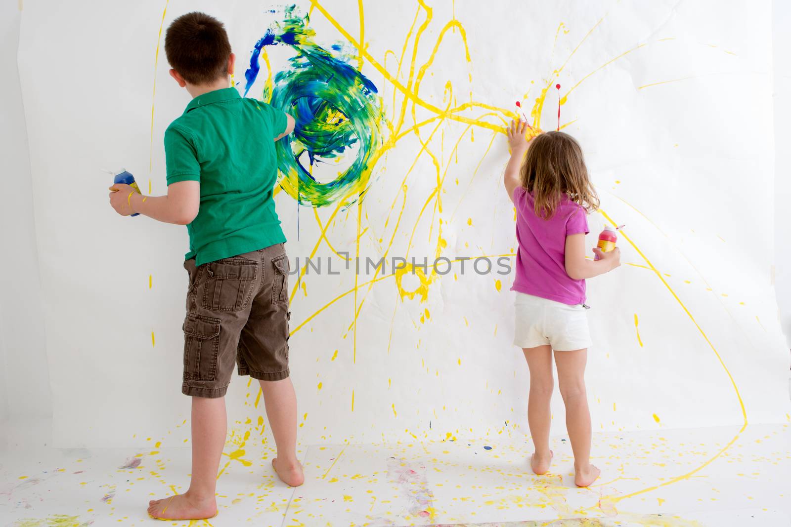 Two young children, a ten year old boy and three year old girl, freehand painting on a wall with colorful acrylic paints creating an abstract design viewed from behind