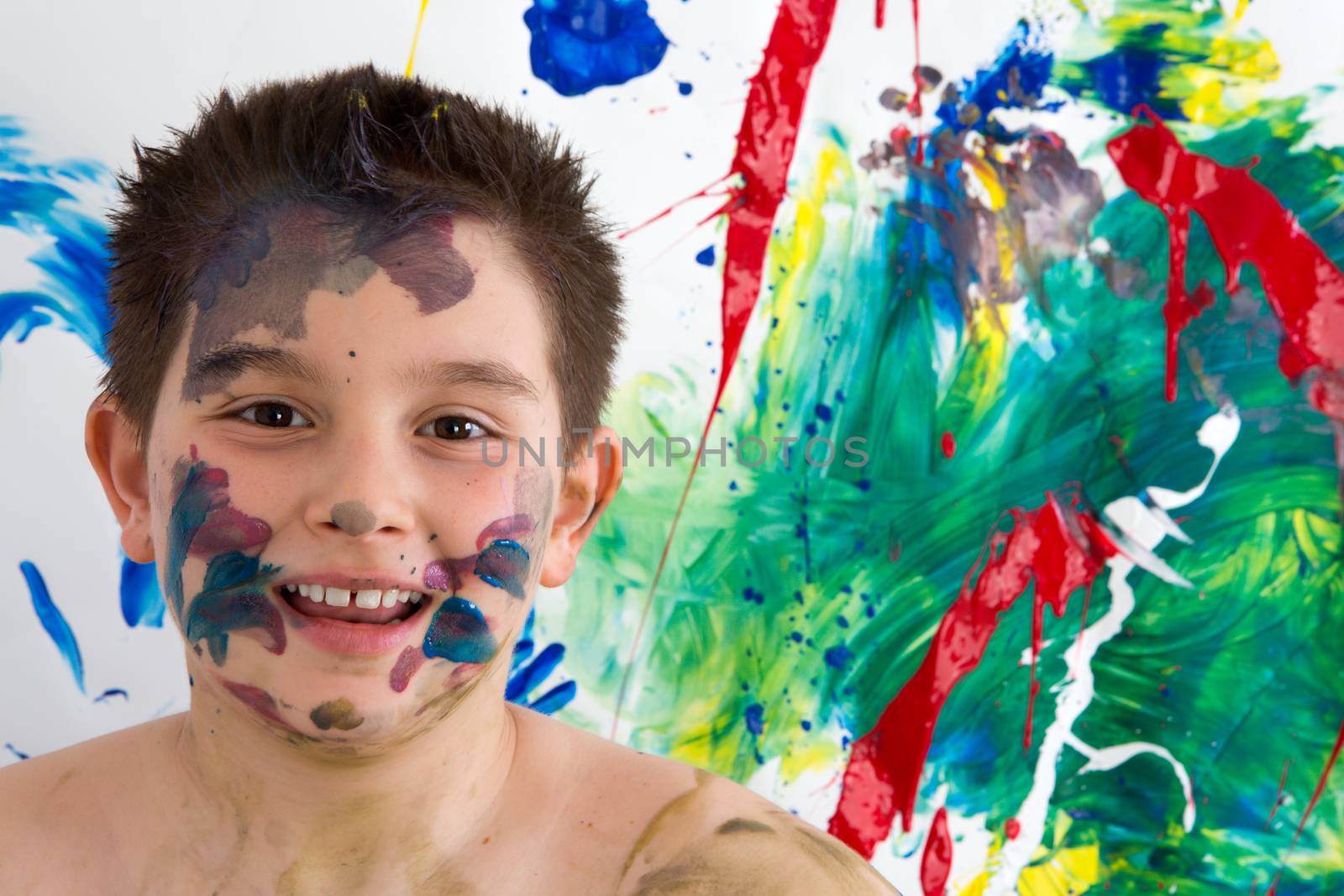 Happy creative little boy with paint splodges on his face standing in front of his bright colorful modern abstract painting grinning at the camera