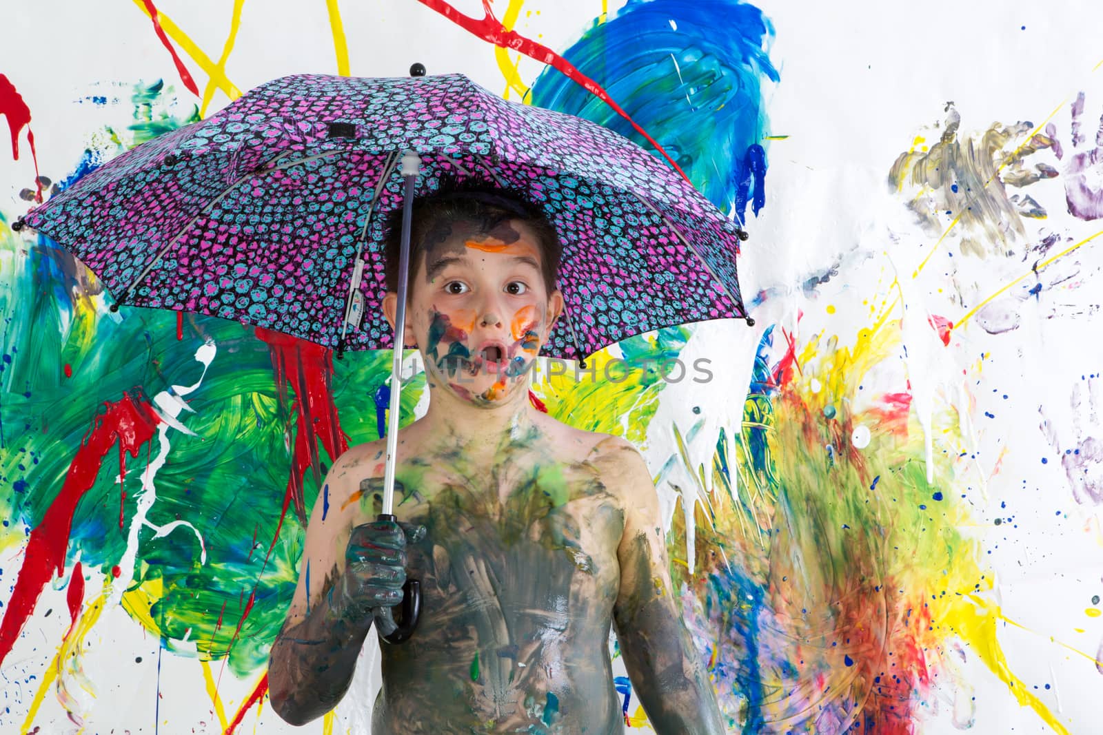 Cute funny shirtless little boy covered in fresh paint standing under an umbrella in front of his vibrant colorful contemporary artwork looking at the camera with an astonished look