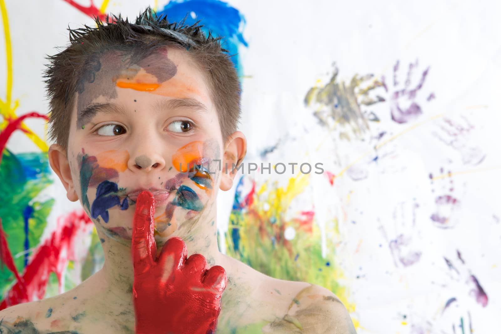 Thoughtful creative little boy covered in paint standing with his finger to his lips looking contemplatively to the side in front of his contemporary artwork as he seeks inspiration