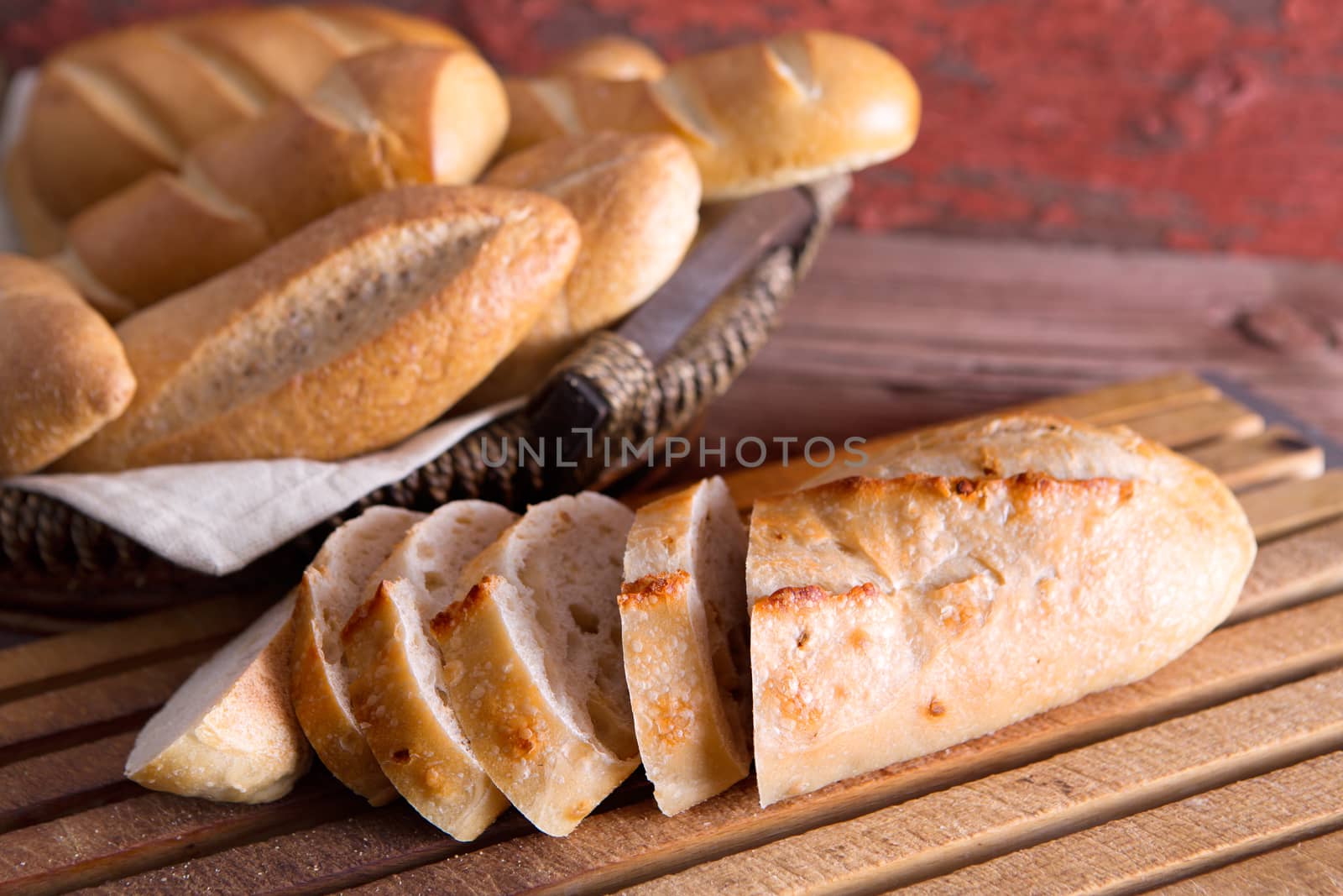 Sliced fresh baguette on a cutting board with a basket of crusty golden rolls and buns on display at a buffet or bakery, close up view