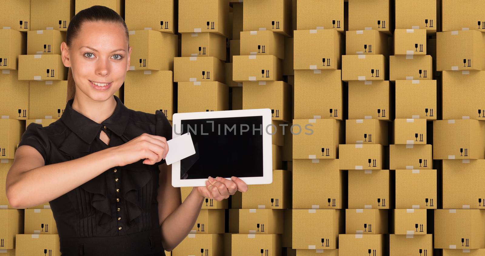 Smiling young woman holging tablet and blank card and looking at camera on abstract cardboard boxes set  background