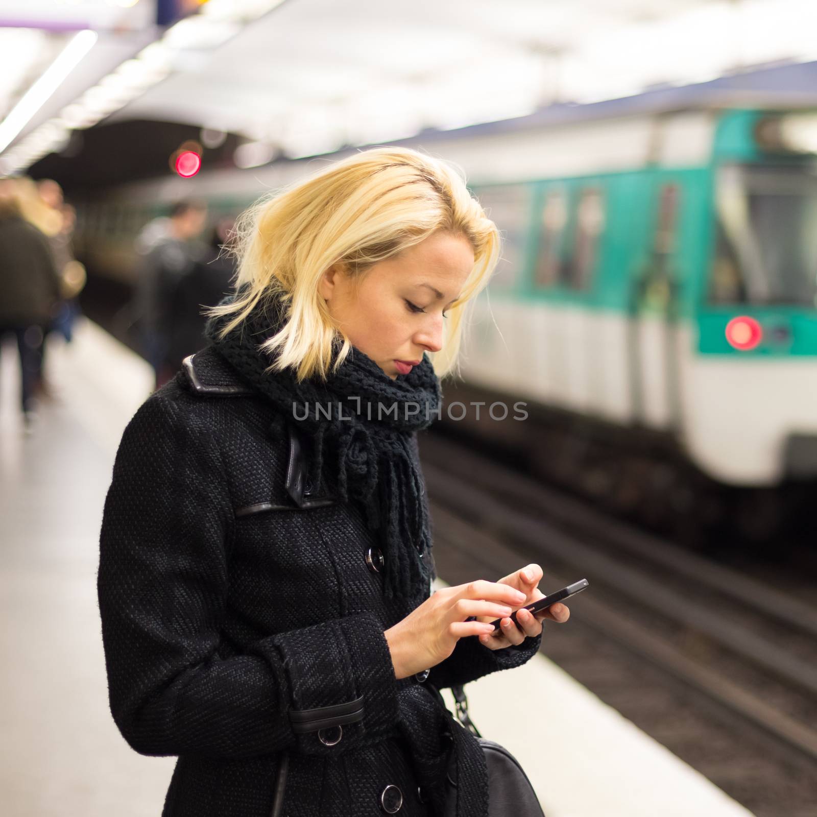 Young woman in winter coat with a cell phone in her hand waiting on the platform of a railway station for train to arrive. Public transport.  