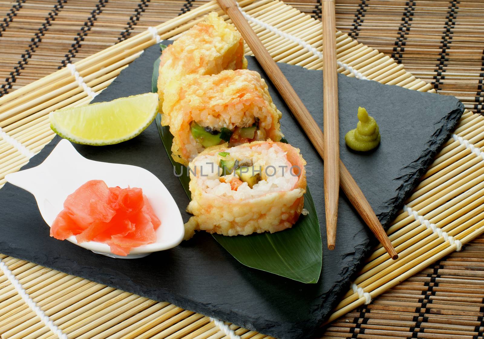 Hot Tempura Roll with Salmon and Avocado Served with Lime and Ginger on Black Plate closeup on Straw Mat background