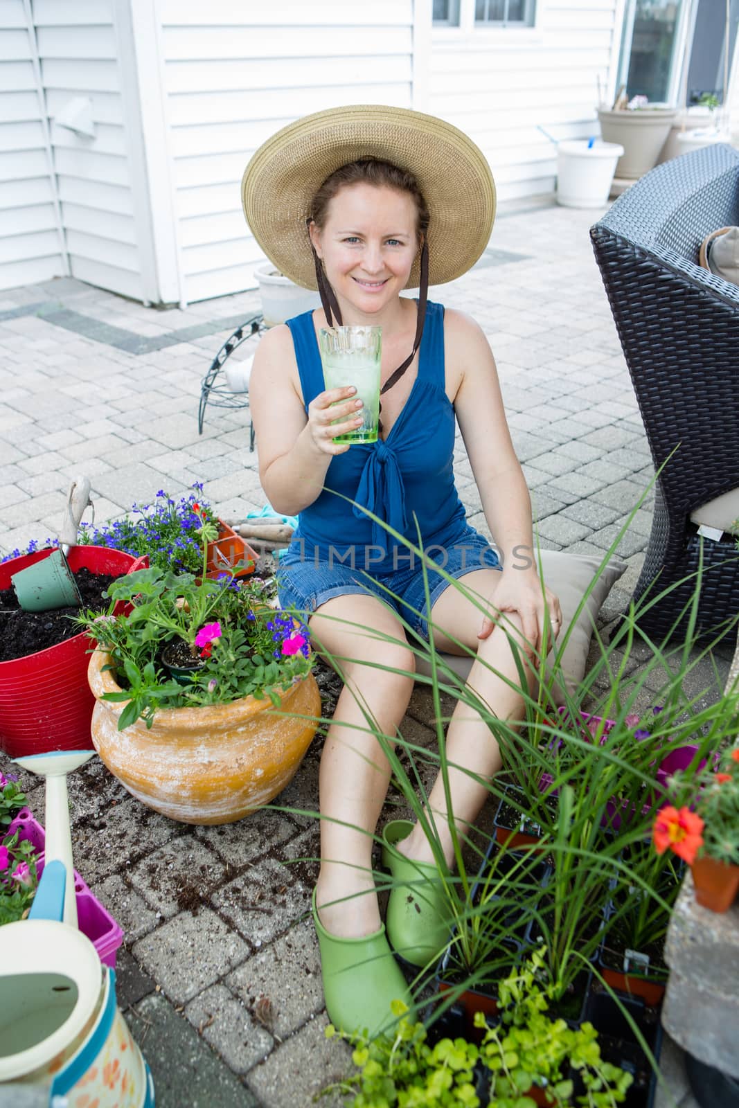 Attractive woman gardener wearing a straw sunhat sitting on her patio enjoying a refreshing iced drink as she takes a break from potting up houseplants from the nursery in springtime