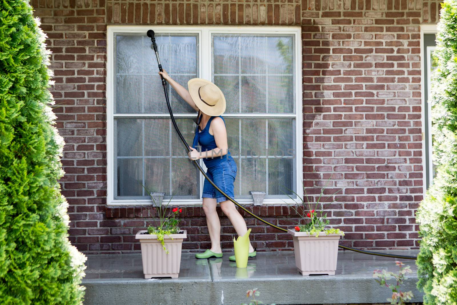 Housewife standing on a patio washing the windows of her house with a hose attachment as she spring-cleans the exterior at the start of the new spring season