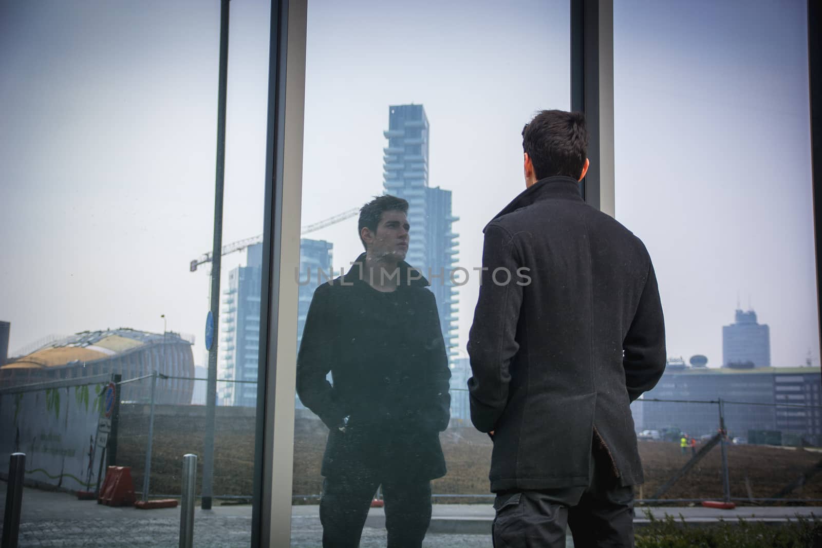 Stylish trendy young man standing outdoor in front of office window, looking at his reflection