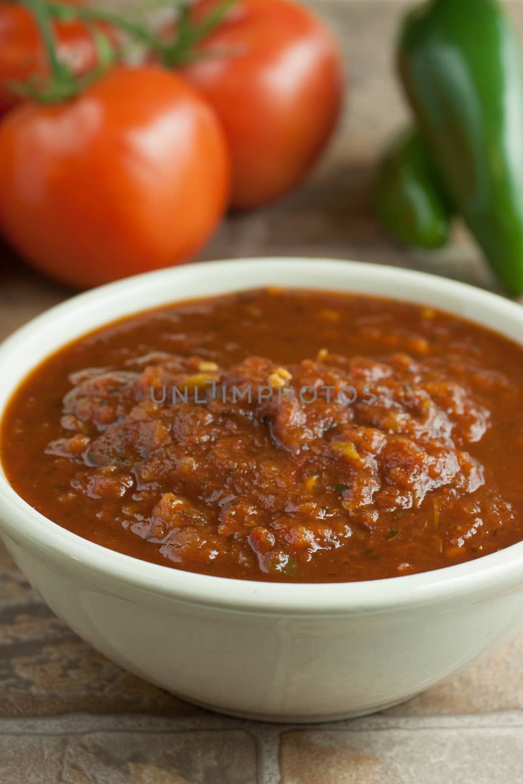 Fresh salsa in a white bowl on a tile surface with tomatoes and jalapenos in the background.