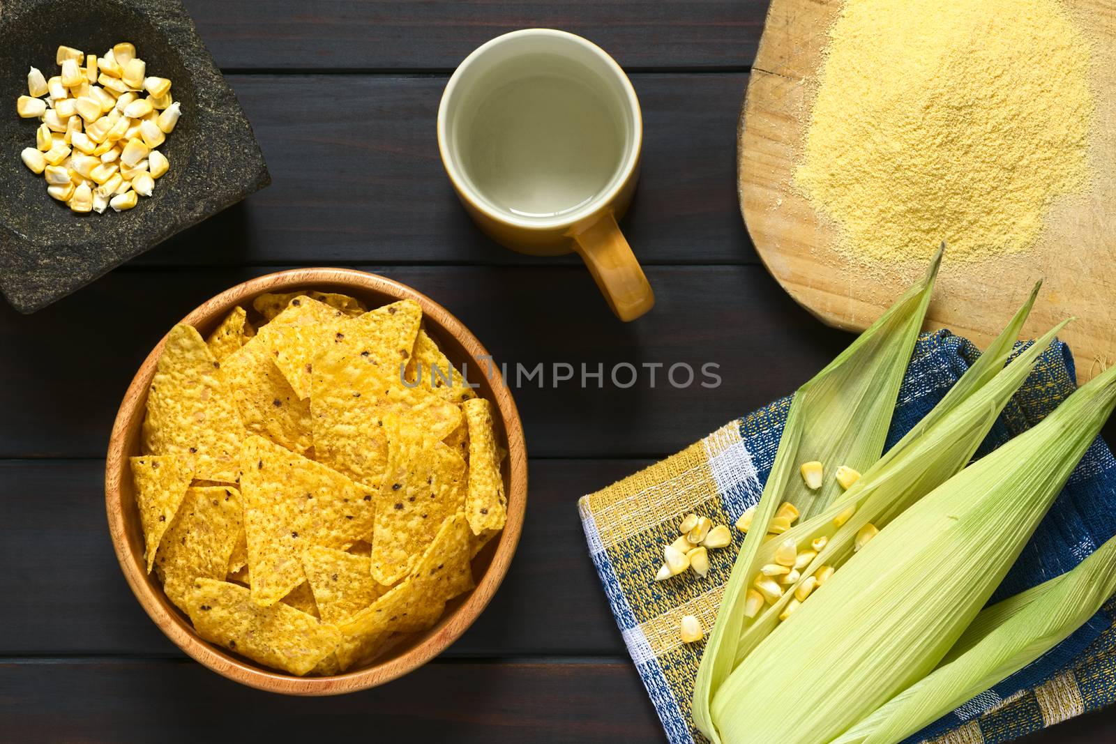 Overhead shot of tortilla chips in wooden bowl surrounded by its ingredients water, cornmeal and corn, photographed on dark wood with natural light