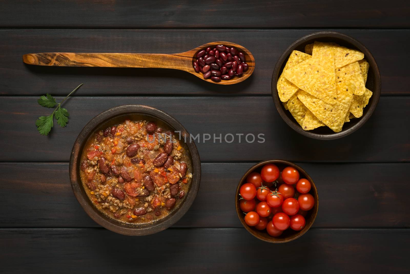 Overhead shot of chili con carne and tortilla chips with ingredients dried kidney beans and cherry tomatoes, photographed on dark wood with natural light