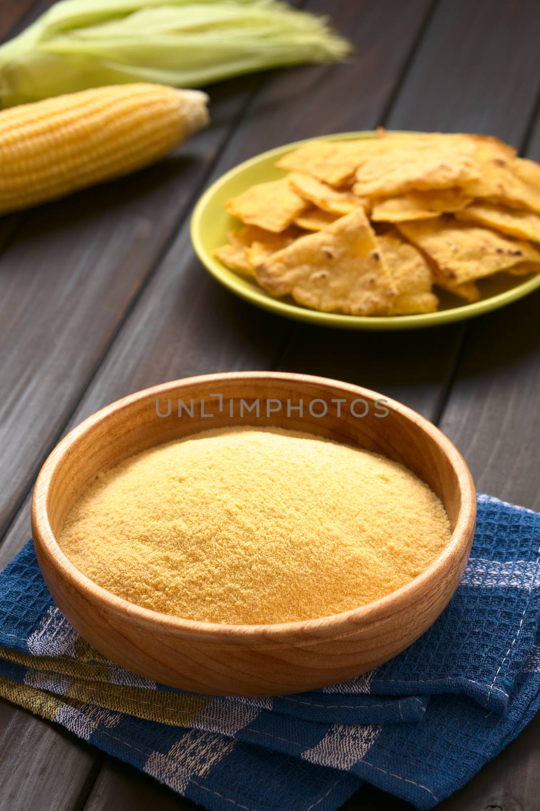 Wooden bowl of cornmeal with homemade tortilla chips and cobs of corn in the back, photographed on dark wood with natural light (Selective Focus, Focus one third into the cornmeal)