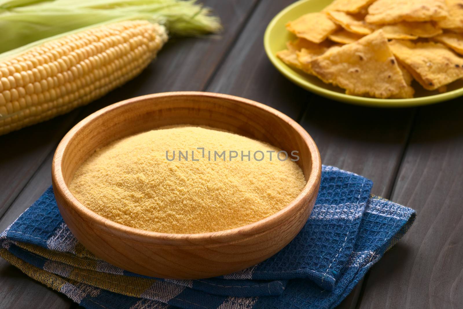 Wooden bowl of cornmeal with homemade tortilla chips and cobs of corn in the back, photographed on dark wood with natural light (Selective Focus, Focus one third into the cornmeal)