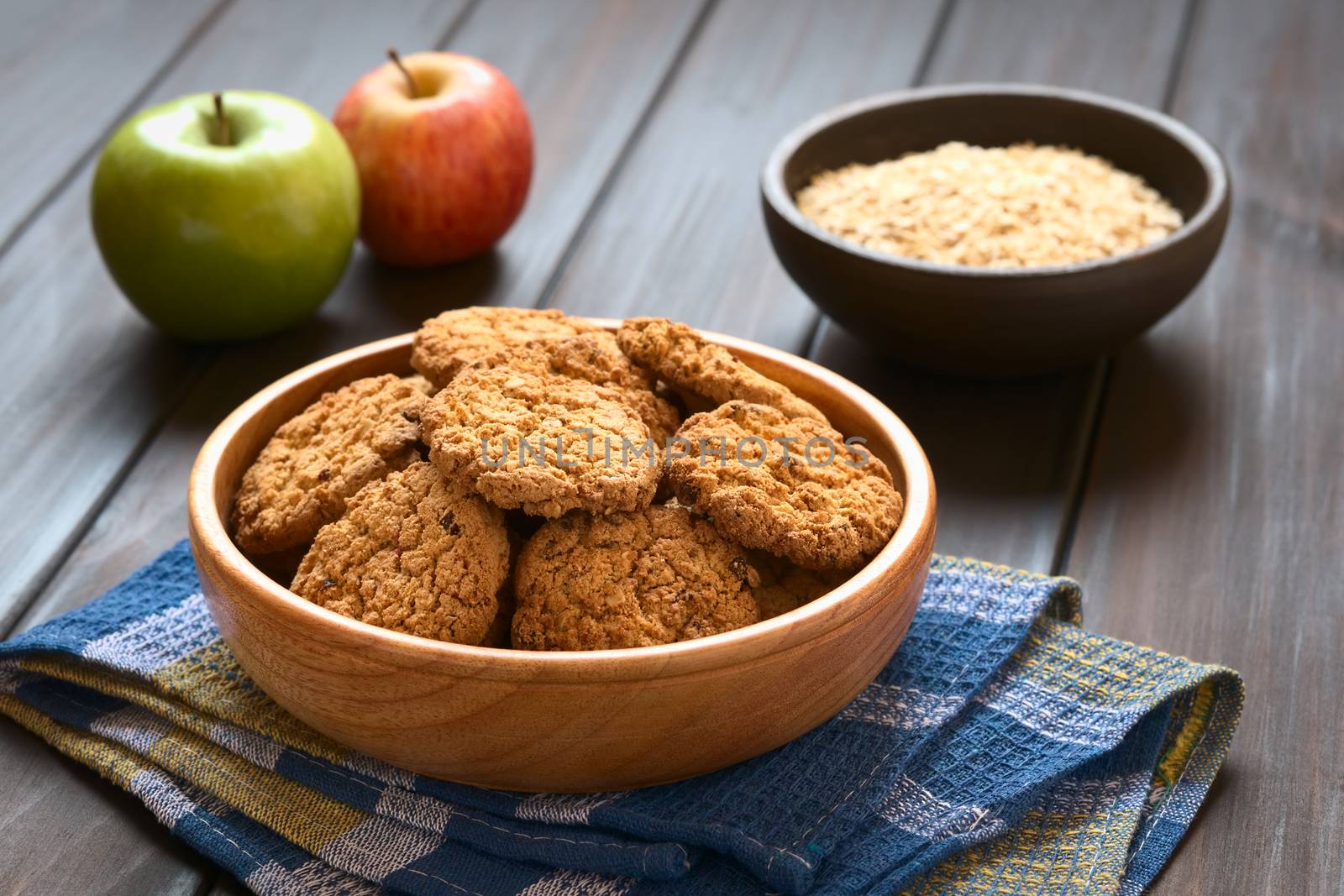 Oatmeal and apple cookies in wooden bowl with apples and oatmeal in the back, photographed with natural light (Selective Focus, Focus on the front of the cookie in the middle)
