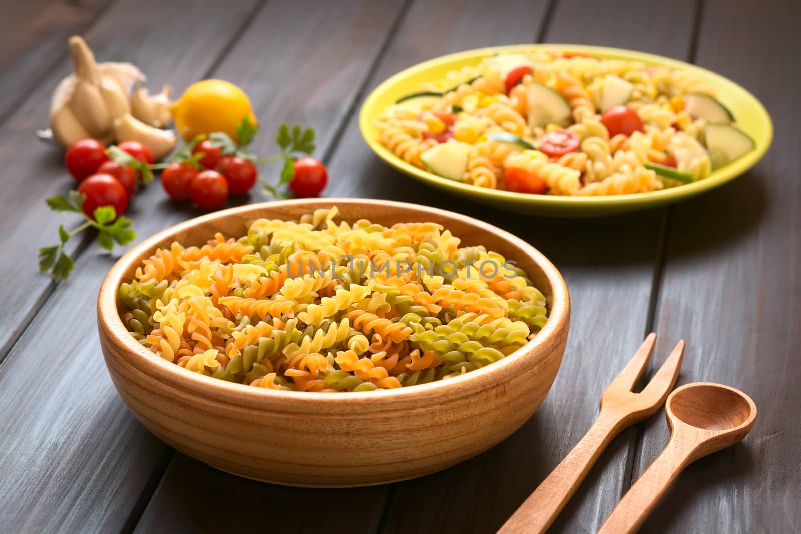 Raw tricolor fusilli or rotini pasta (traditional, tomato and spinach taste) in wooden bowl with pasta salad and ingredients in the back, photographed with natural light (Selective Focus, Focus one third into the pasta) 