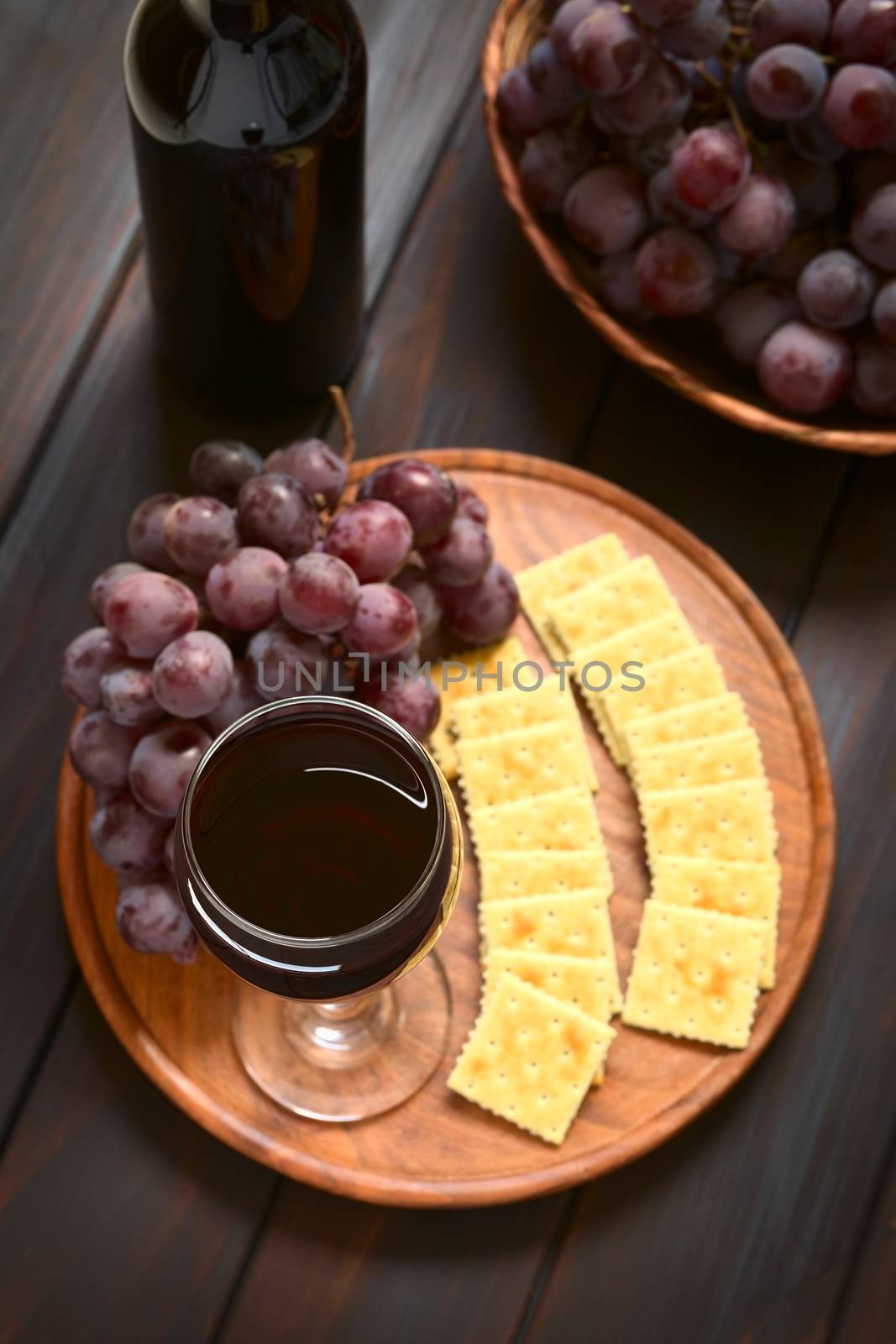 Glass of red wine with red globe grape, crackers and a bottle of wine, photographed on dark wood with natural light (Selective Focus, Focus on the rim of the wine glass)