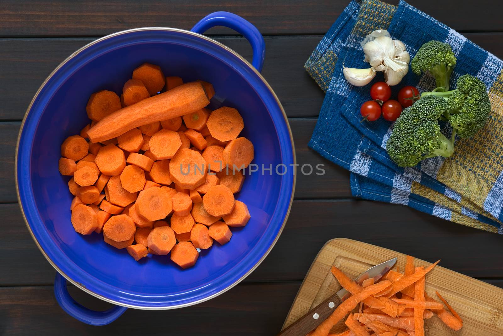 Overhead shot of fresh raw sliced carrot in blue metal strainer with broccoli, cherry tomato, garlic and carrot peels on the side, photographed on dark wood with natural light