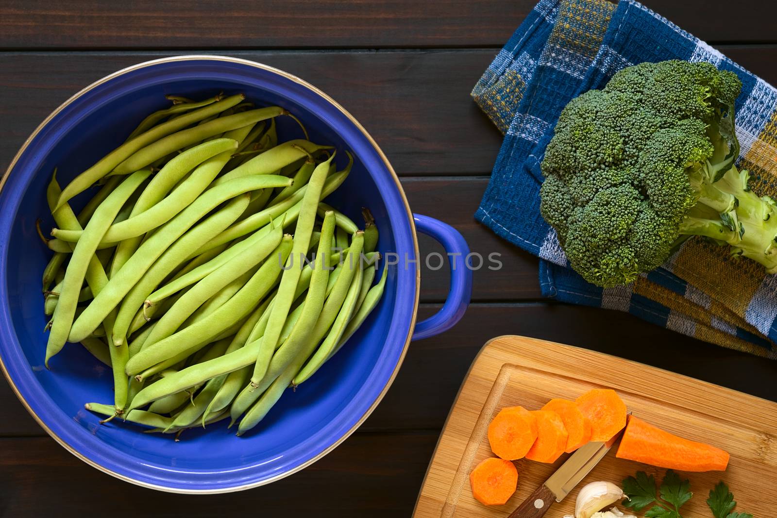 Overhead shot of green beans in blue metal strainer with broccoli, and cut carrot on the side, photographed on dark wood with natural light