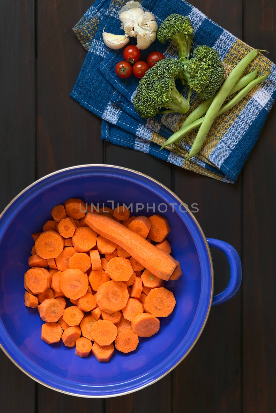 Overhead shot of fresh raw sliced carrot in blue metal strainer with broccoli, cherry tomato, garlic, green bean on kitchen towel, photographed on dark wood with natural light