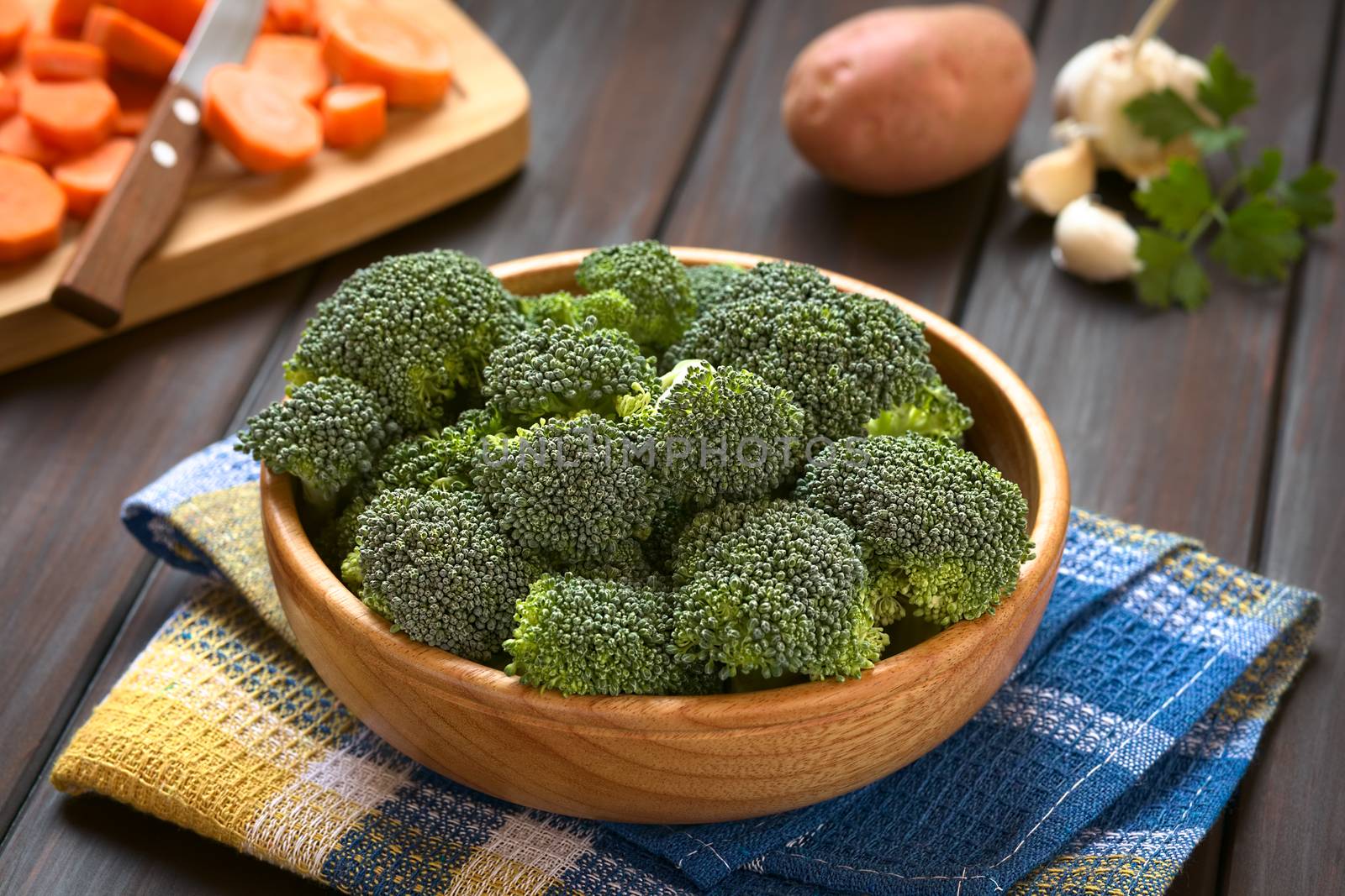Fresh raw broccoli florets in wooden bowl with carrot slices on wooden board, potato, garlic and parsley in the back, photographed on dark wood with natural light (Selective Focus, Focus one third into the broccoli)