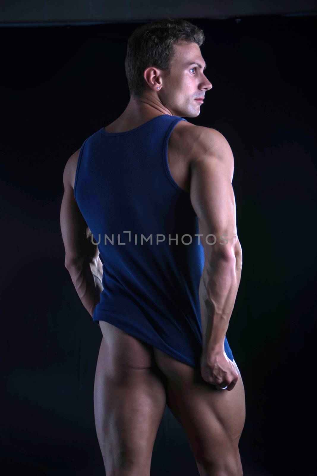 Back of young athletic man pulling down tanktop on ripped muscular torso, isolated on dark background