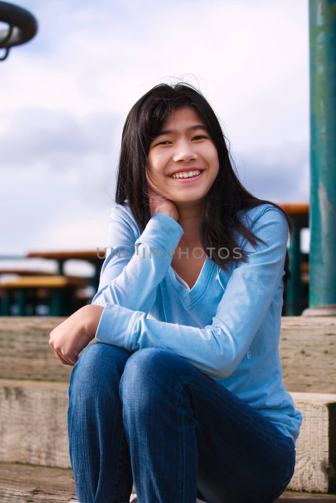 Young biracial teen girl in blue shirt and jeans sitting on wooden steps outdoors on overcast cloudy day