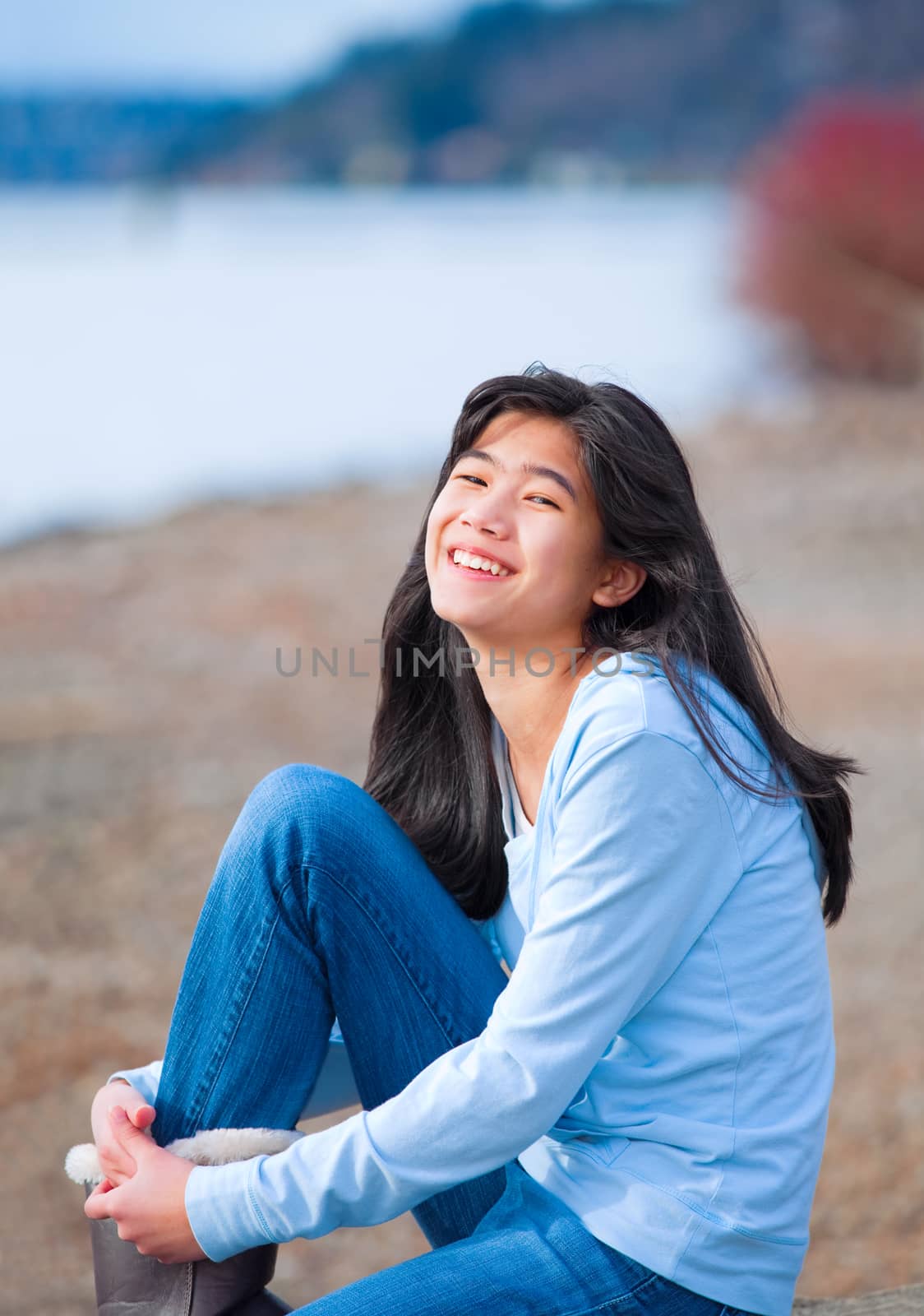 Young biracial teen girl in blue shirt and jeans sitting along rocky lake shore on bright overcast day outdoors