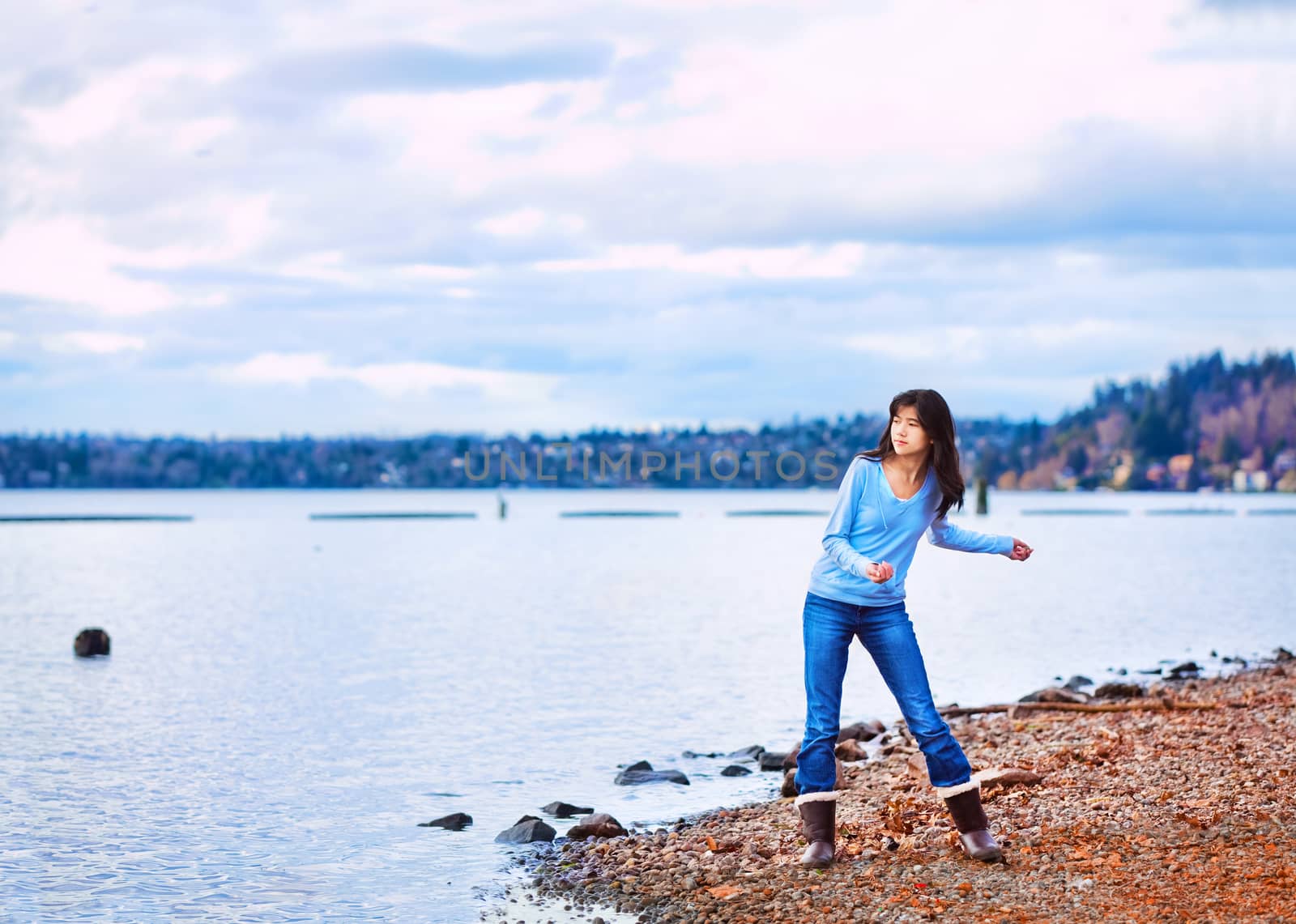 Young biracial teen girl in blue shirt and jeans along rocky lake shore, throwing rocks into the water on cloudy overcast day