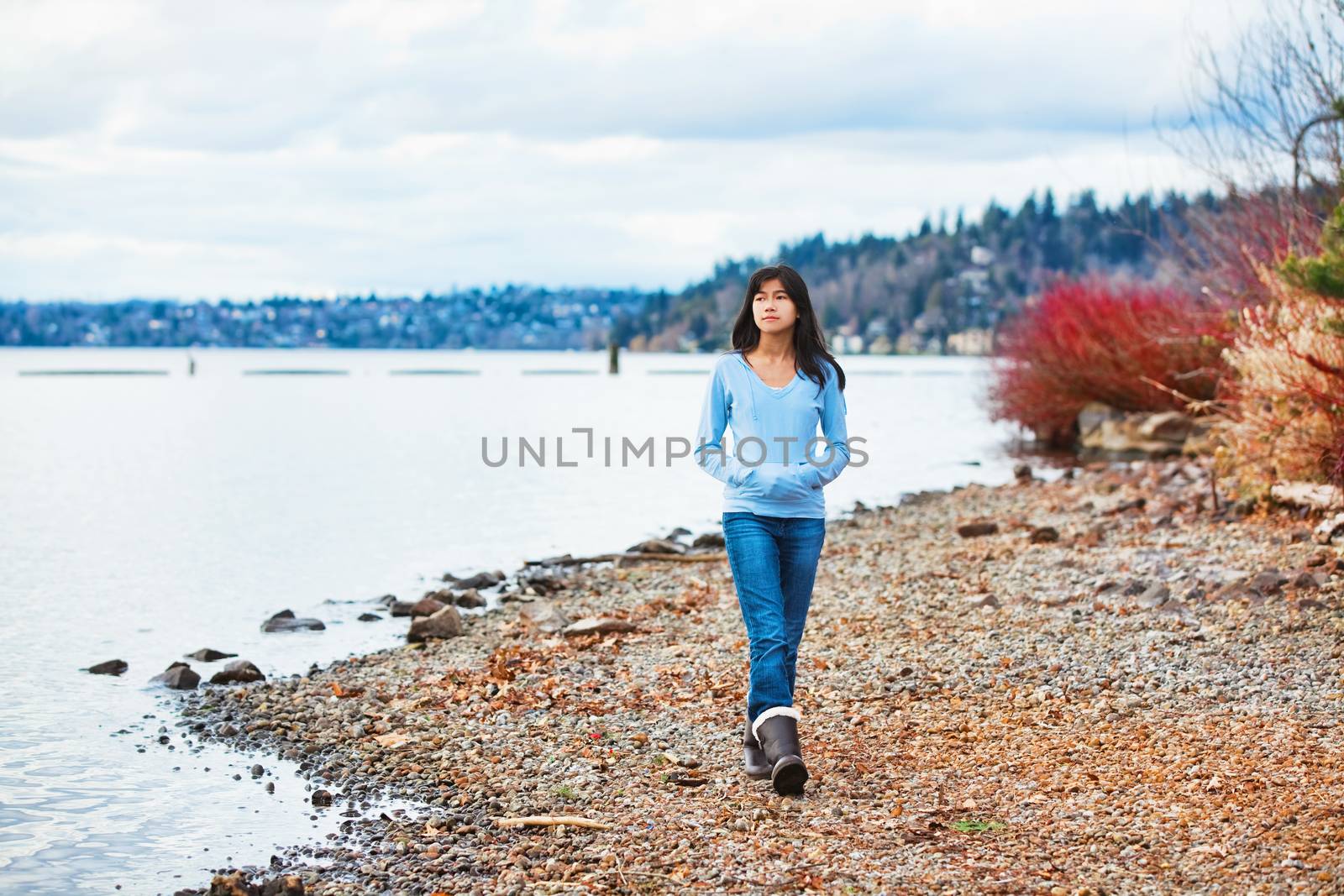 Young biracial teen girl in blue shirt and jeans walking along rocky shoreline of lake in early spring or fall