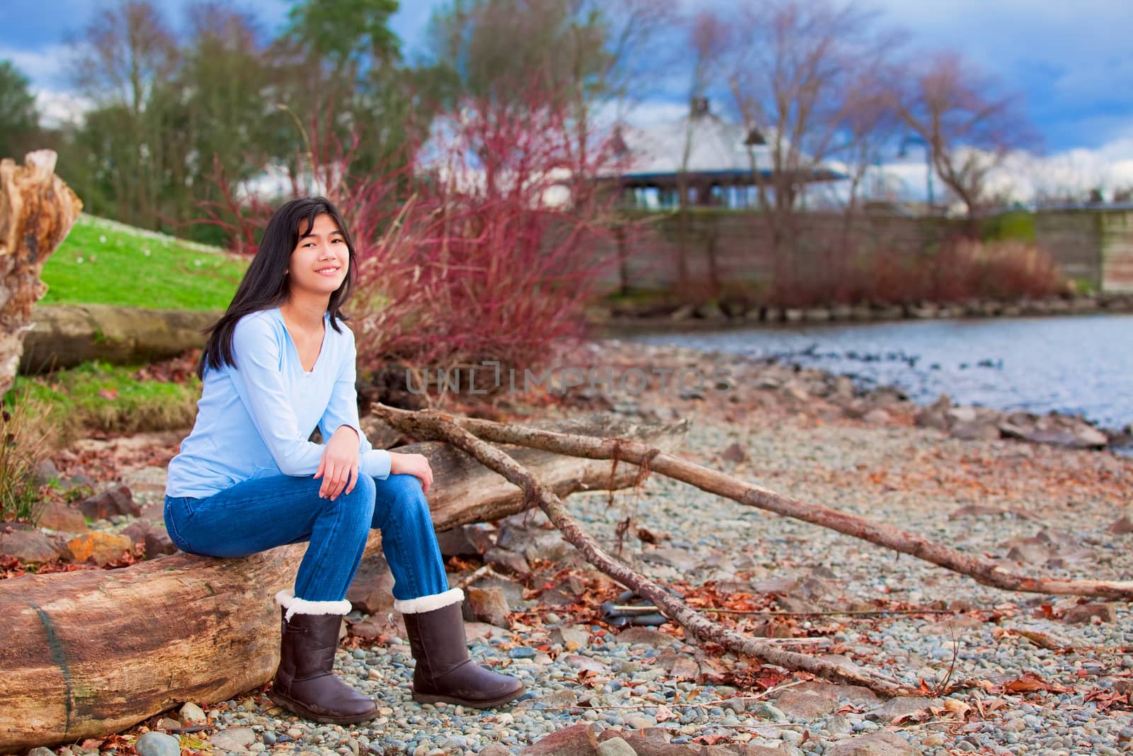 Young teen girl sitting on log along rocky beach of lake by jarenwicklund