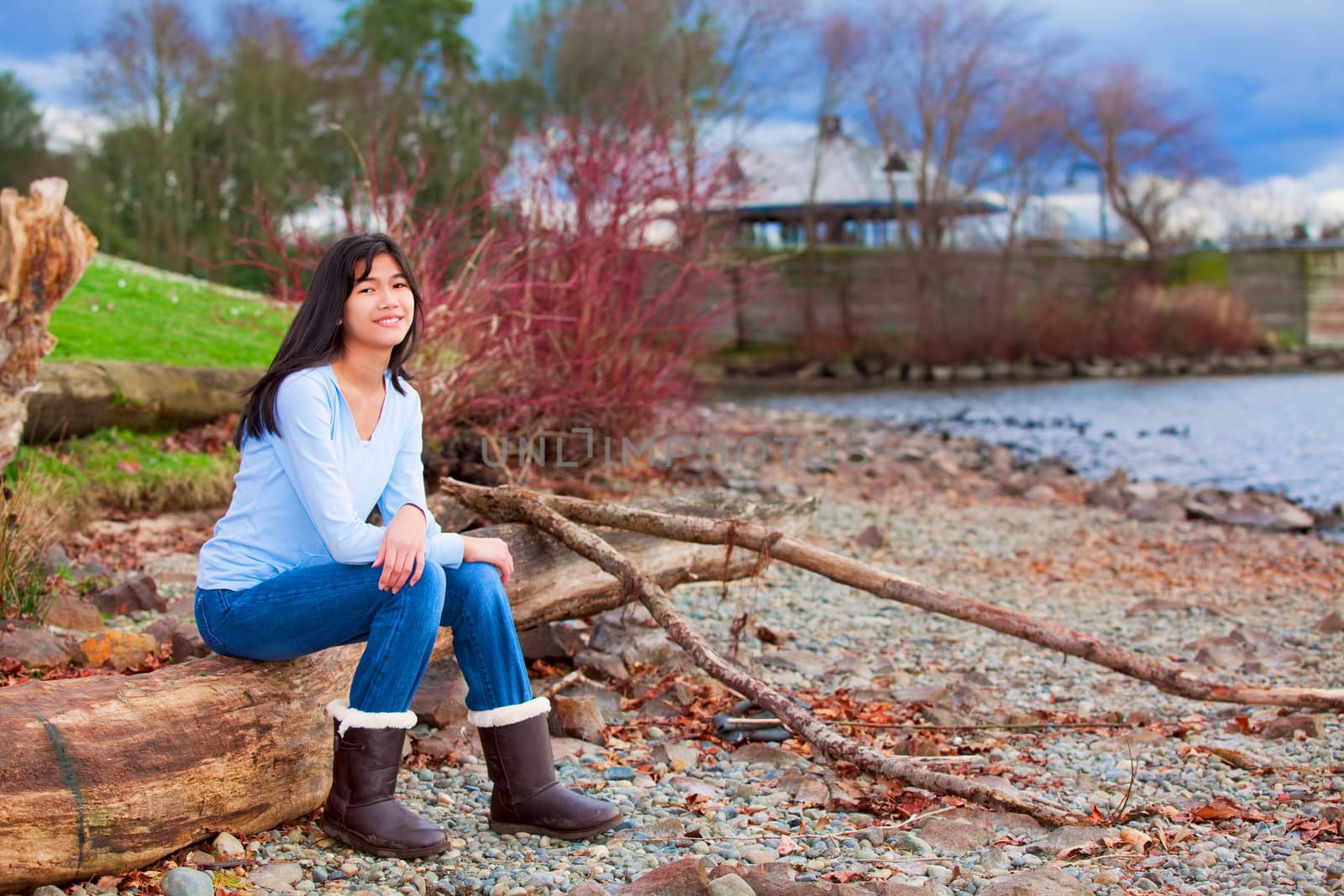 Young biracial teen girl in blue shirt and jeans sitting on large log on rocky beach by lake