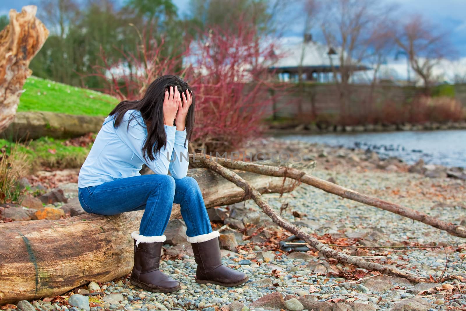 Sad young biracial teen girl in blue shirt and jeans sitting on log along rocky beach by lake