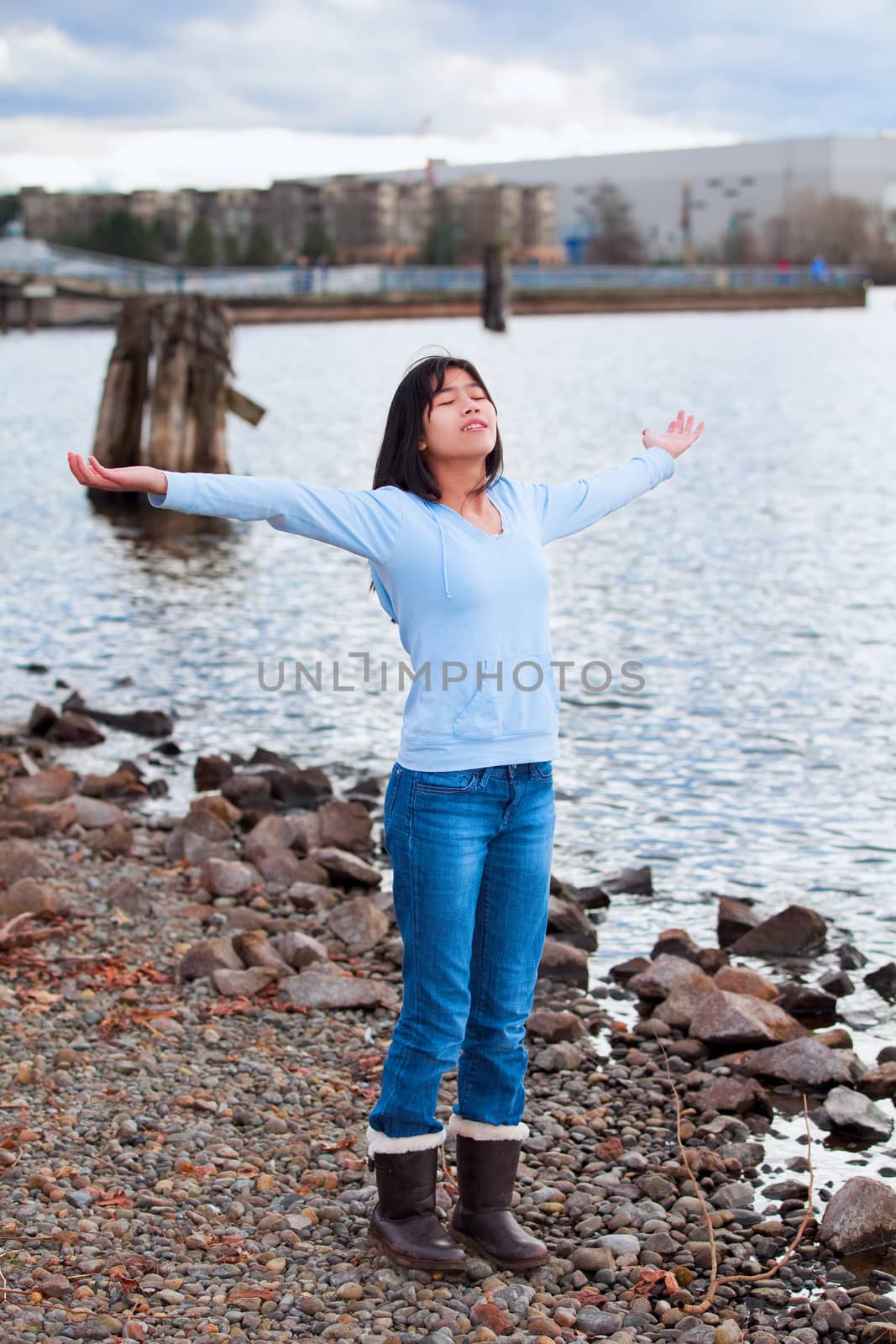 Young biracial teen girl in blue shirt and jeans, arms lifted and outstretched, praising God on rocky shore by lake on cloudy day