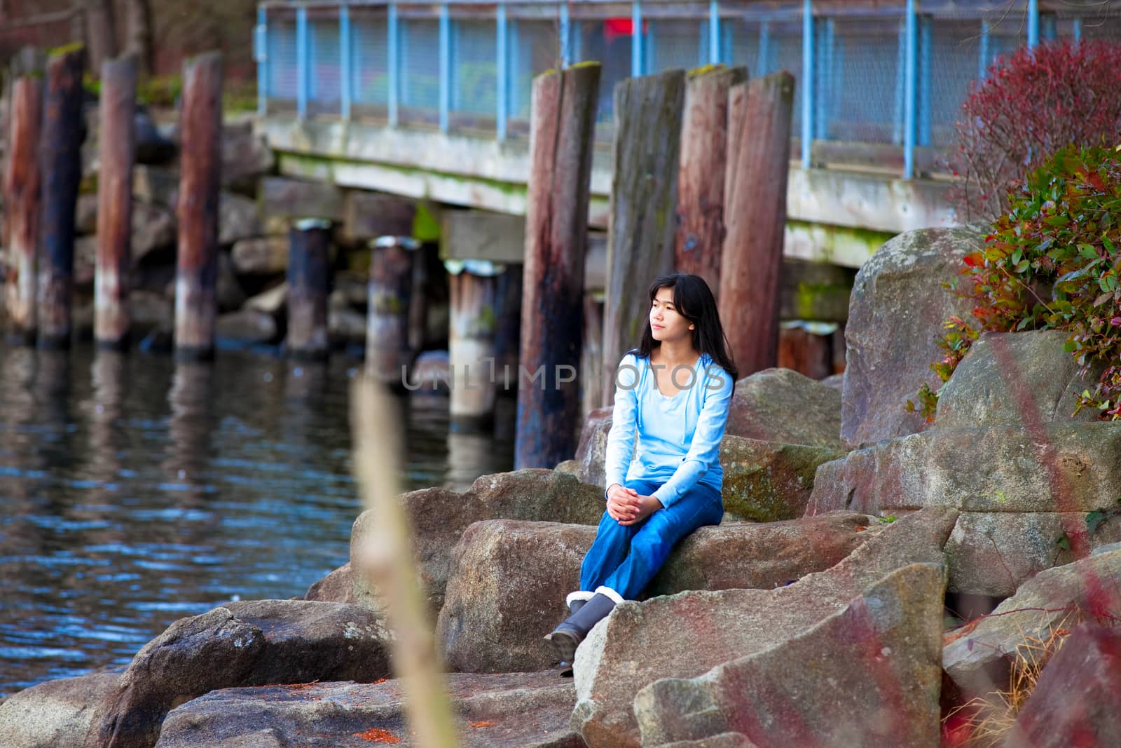 Young biracial teen girl in blue shirt and jeans sitting on large boulders along lake shore, looking out over water