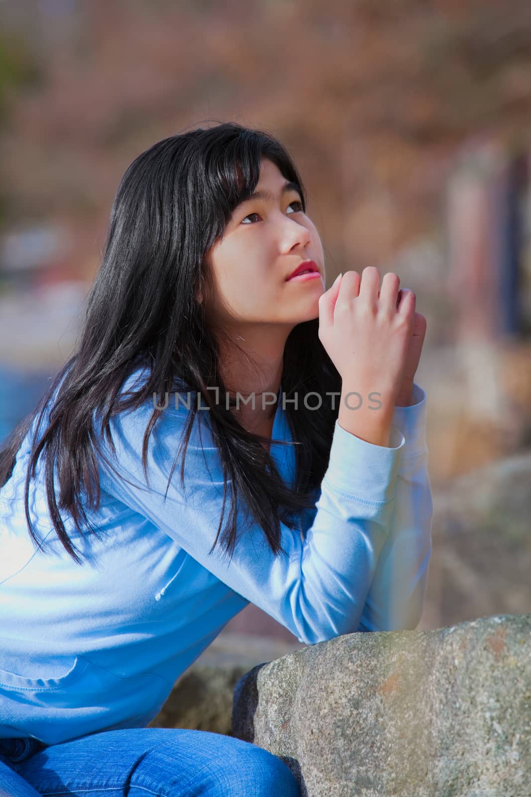 Young biracial teen girl in blue shirt and jeans quietly sitting outdoors leaning on rocks praying