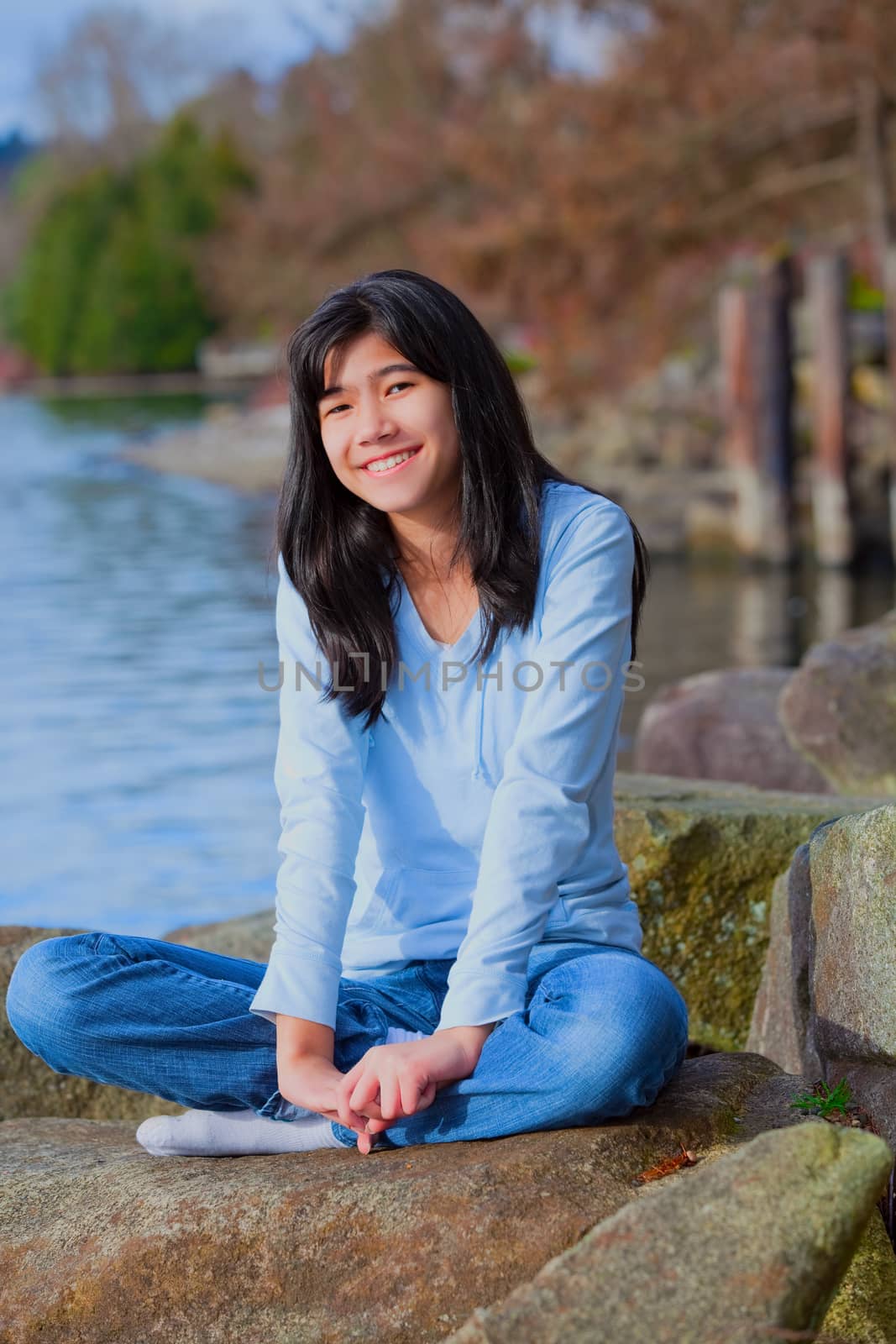 Young biracial teen girl in blue shirt and jeans sitting on large boulder or rocks along rocky lake shore, smiling and reclining