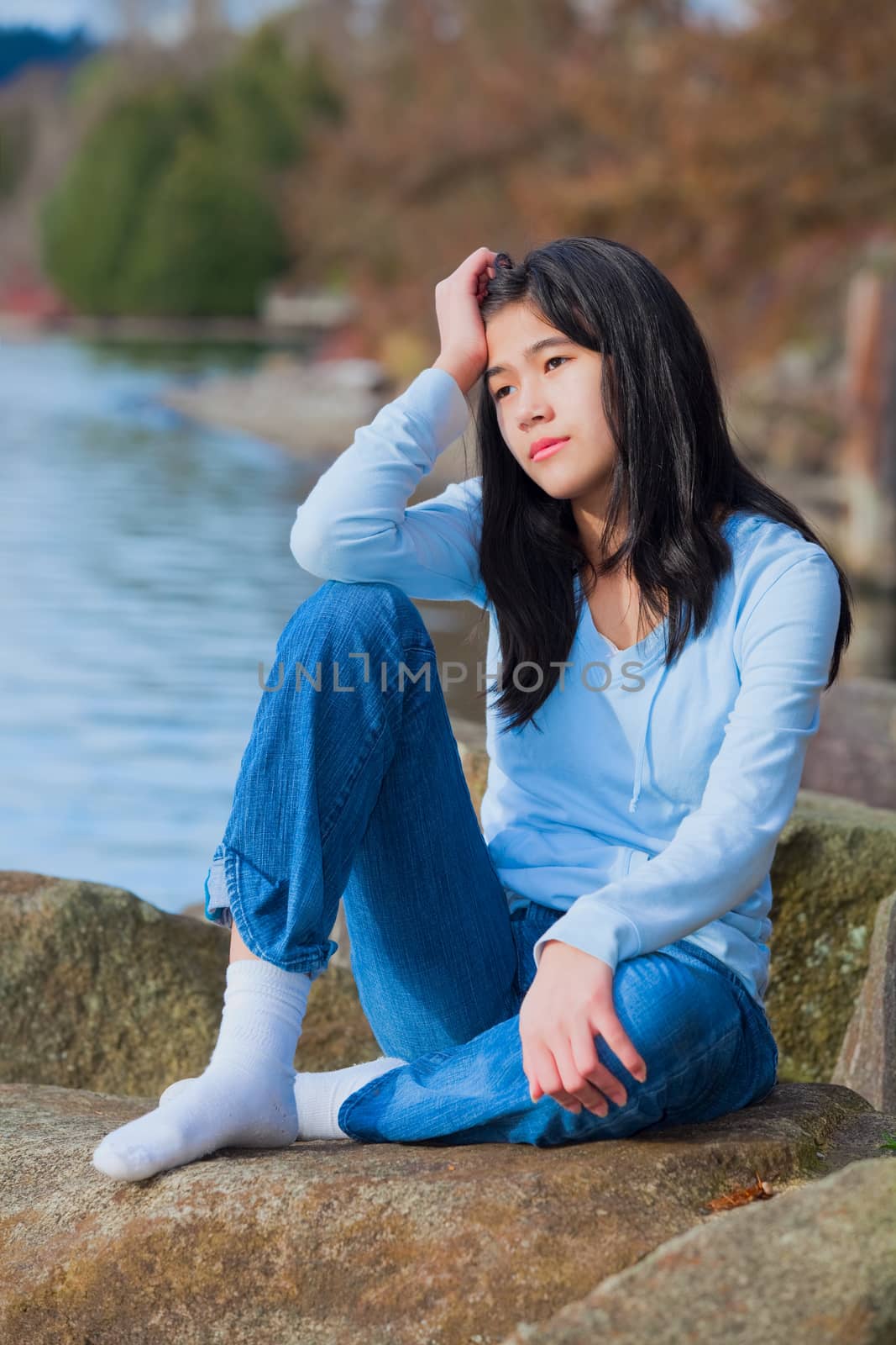 Young unhappy biracial teen girl in blue shirt and jeans sitting on rocks along lake shore, looking off to side, resting head in hand and one knee raised.