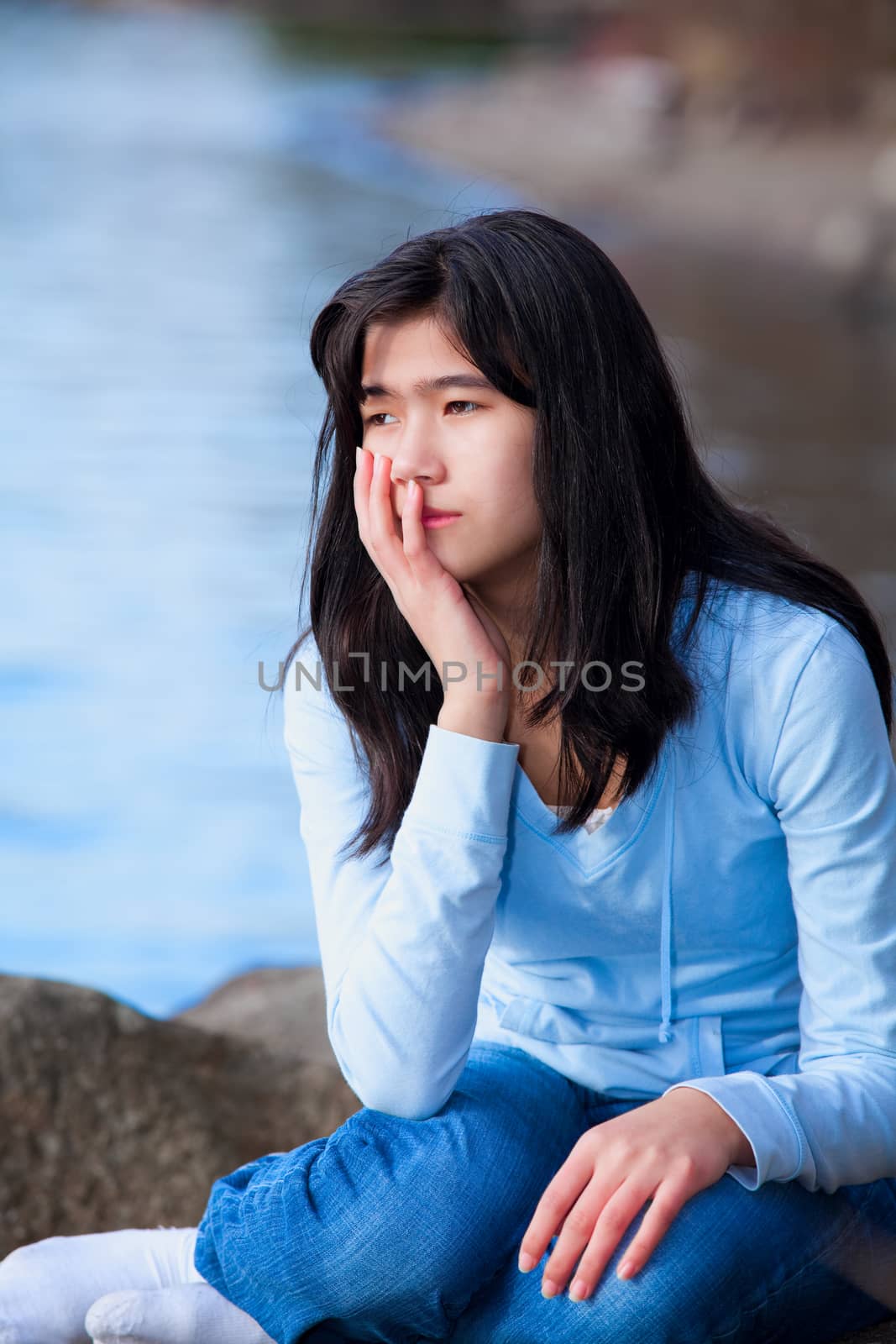 Sad teen girl sitting on rocks along lake shore, lonely expressi by jarenwicklund