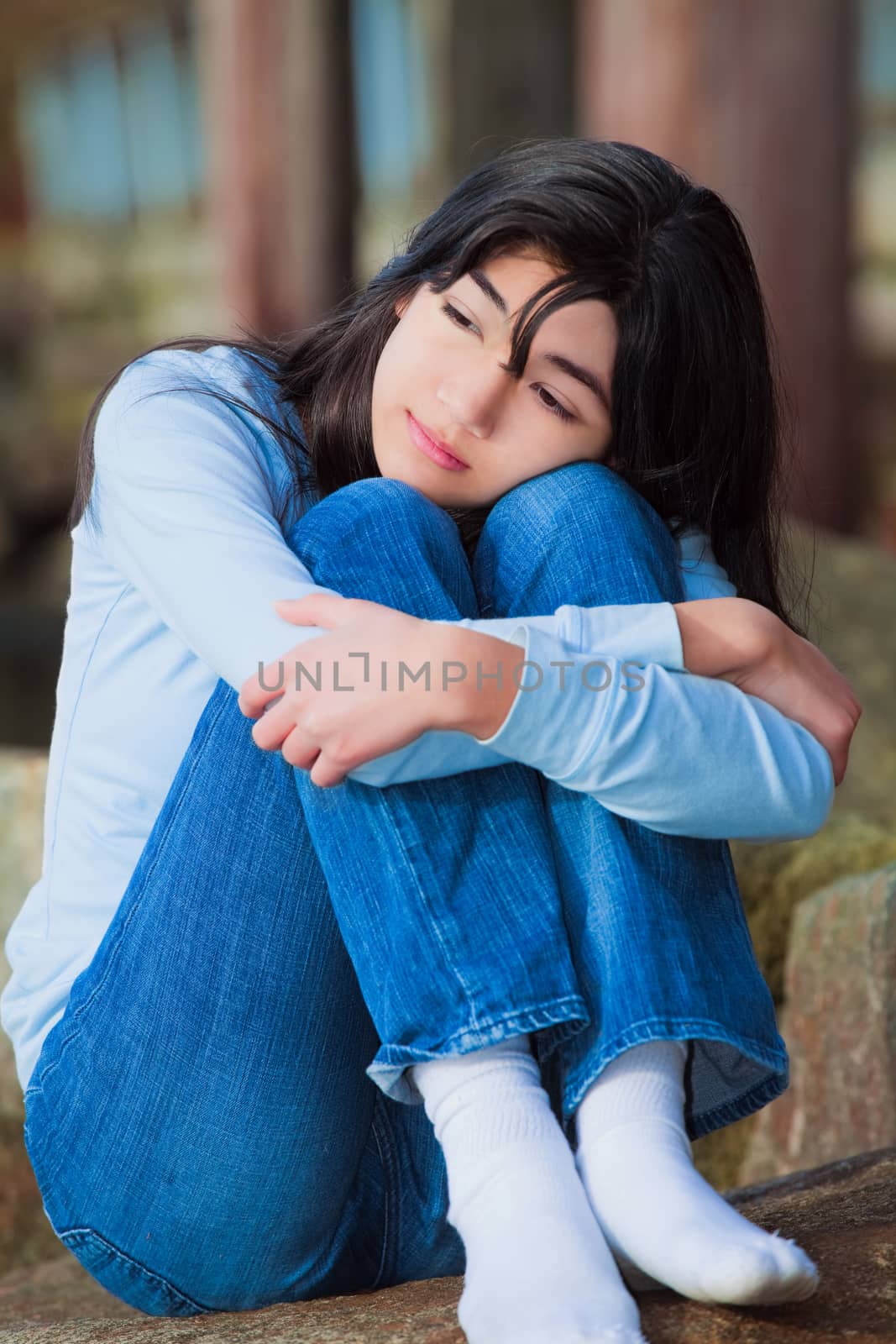 Sad teen girl sitting on rocks along lake shore, lonely expressi by jarenwicklund