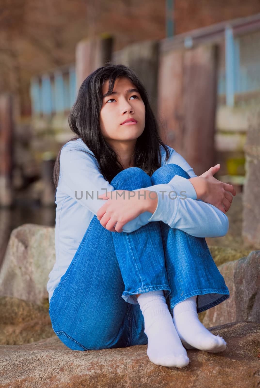 Sad biracial teen girl in blue shirt and jeans sitting on rocks along lake shore, lonely expression