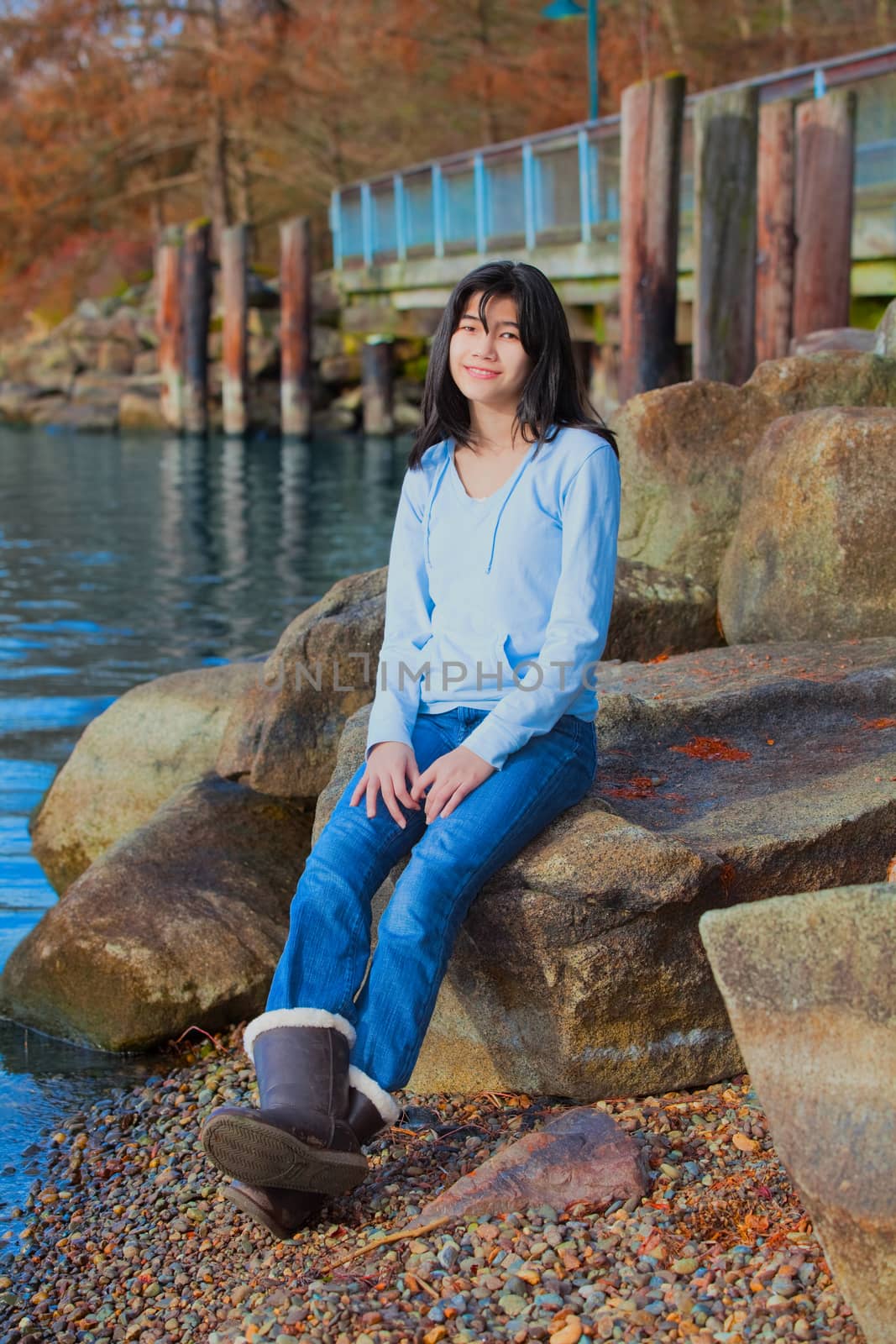 Young biracial teen girl in blue shirt and jeans sitting on large boulder or rocks along rocky lake shore, smiling and reclining